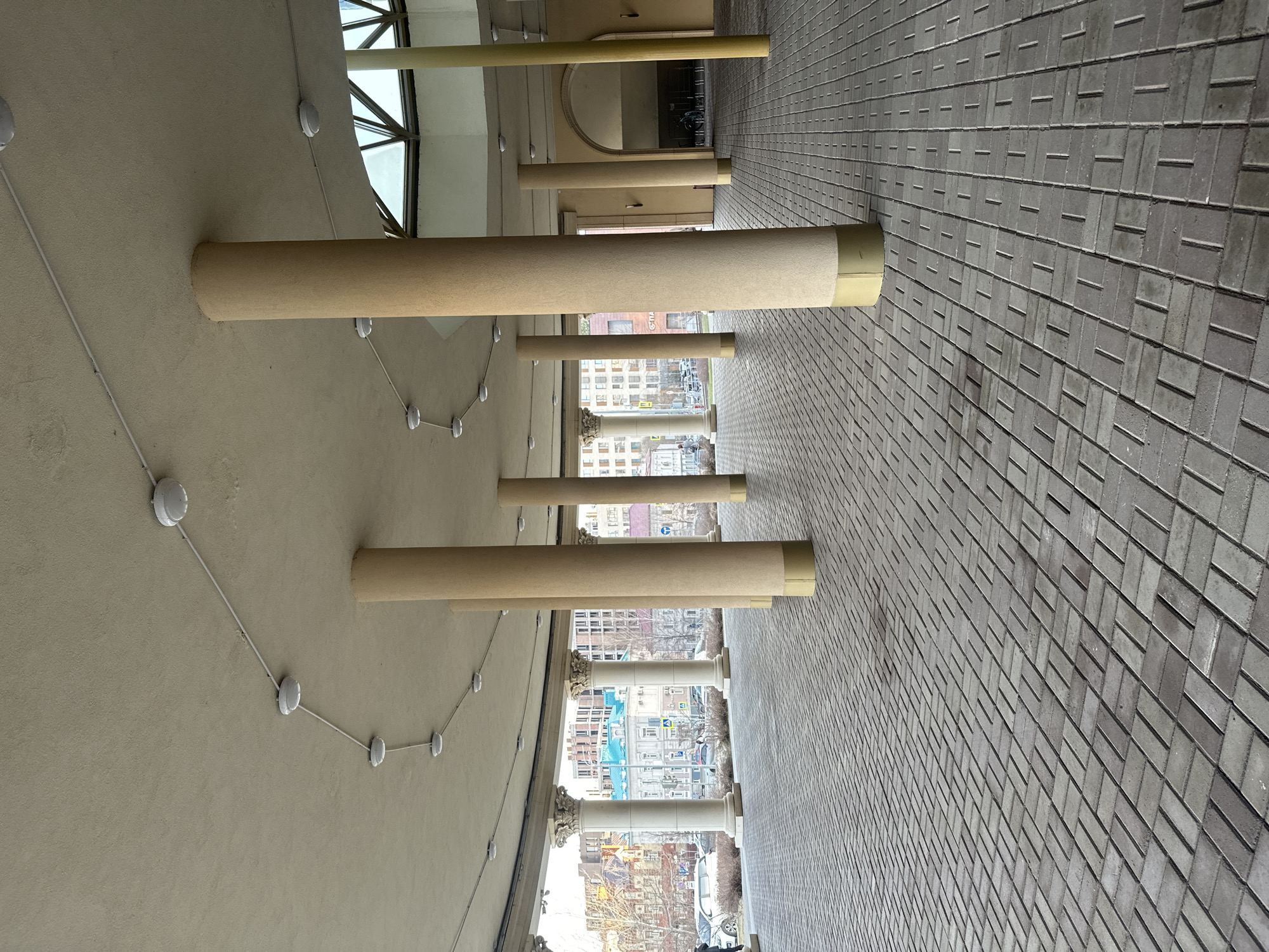 The image shows an architectural detail of a building in Kazan, Russia. It features cylindrical structures protruding horizontally from a cream-colored ceiling or wall, which also displays electrical wiring and ceiling fixtures. On the opposite side, there is a brick-paved surface, likely a sidewalk. Through the spaces between the cylinders, one can see the city landscape, with buildings and some visible traffic. The overall image composition suggests an open-air corridor or arcade.