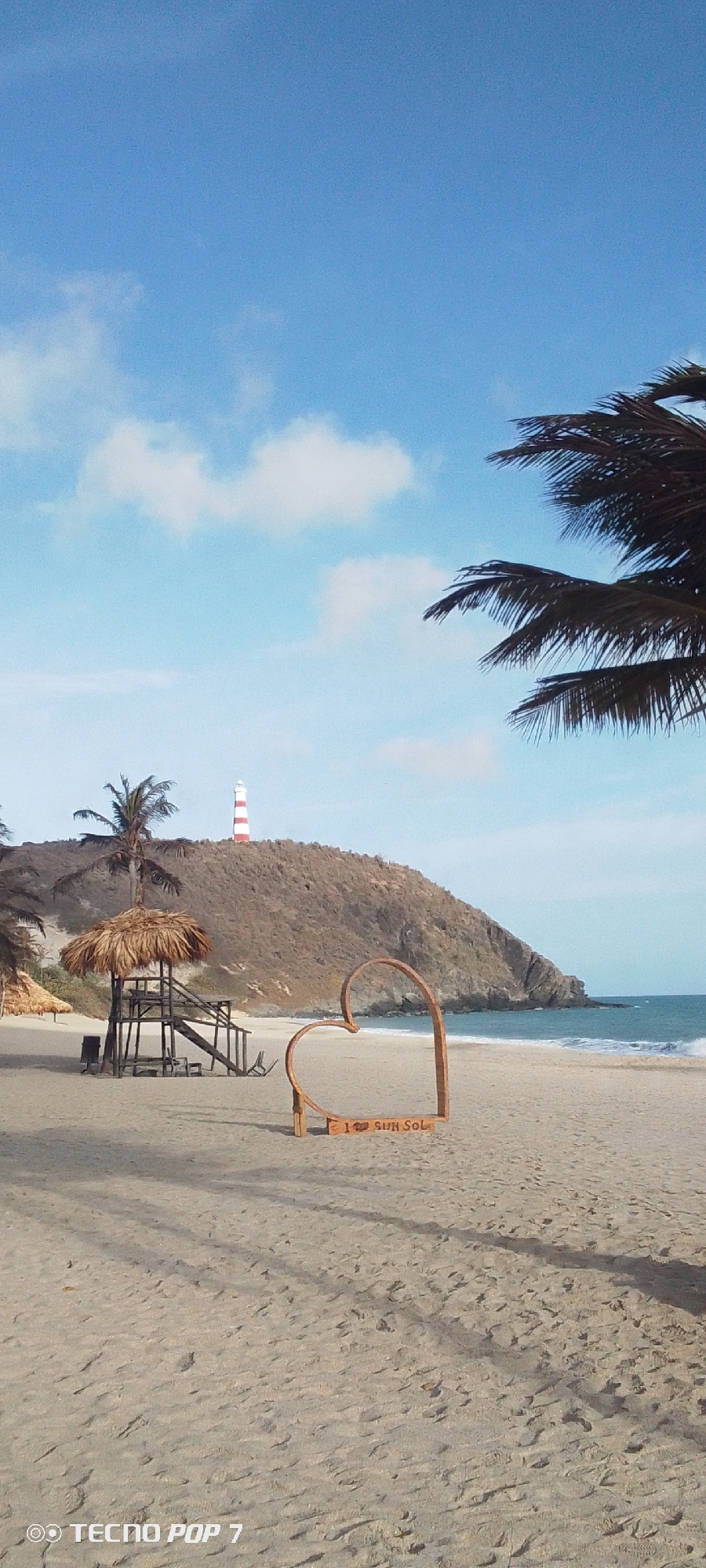 The image captures a beach scene in Pedro González, Venezuela, featuring a sandy beach under a blue sky with scattered clouds. A wooden heart-shaped structure with the inscription "I ❤️ SUN SOL" stands in the foreground. A lifeguard stand with a thatched roof is visible, and in the distance, a hill rises, topped with a red and white striped lighthouse. The ocean is visible at the edge of the beach, and palm trees are seen to the right and left of the frame. Shadows are cast on the sand. The brand "TECNO POP 7" is visible in the bottom left corner.