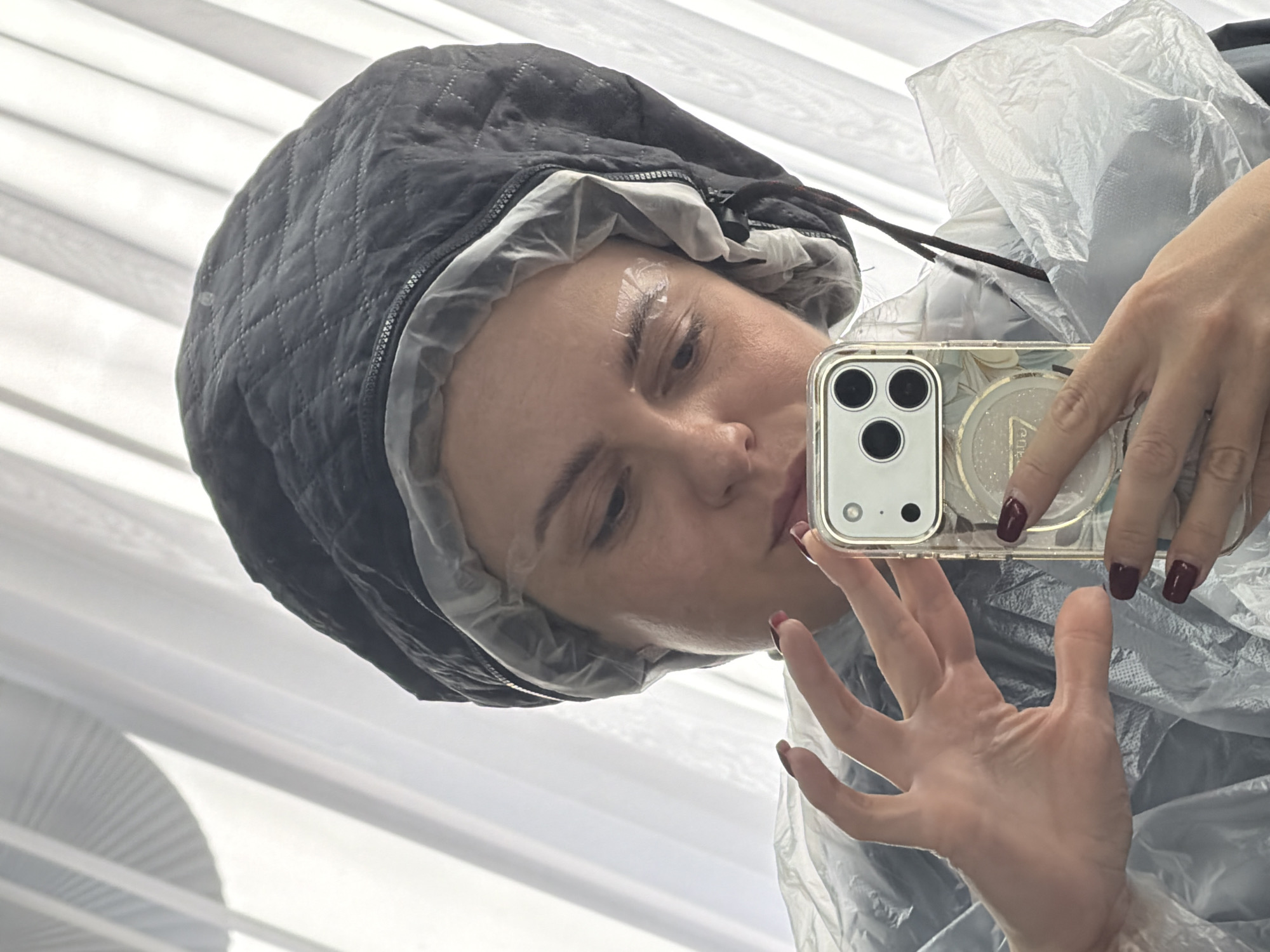 A woman with cream on her eyebrows is taking a selfie. She is wearing a black quilted hood over a plastic shower cap, and a clear plastic smock. She is holding an iPhone in a clear case with a circular phone grip. The background consists of vertical white blinds. The scene is in Votkinsk, Russia.