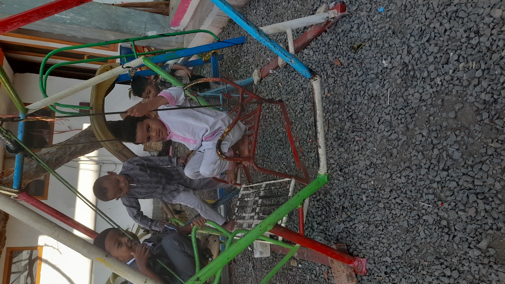 In Selaawi, Indonesia, four young boys are gathered around a colorful, metal swing set on a gravel ground. One boy in the center, wearing a white garment with pink trim, sits on a red swing seat, holding onto the chains. To his left, another boy in a patterned gray shirt and light pants holds a white object, possibly a lollipop, to his mouth. Another boy in a dark gray shirt and pants, wearing a skullcap, is seated to the far left, giving a thumbs-up. A fourth boy, partially visible in the background to the right, wears a plaid shirt. The swing set features red, green, white, and blue painted metal poles, with some of the paint chipped. The ground is covered in small, gray gravel. In the background, a wall with windows and a tree are visible. The overall scene depicts children playing outdoors.