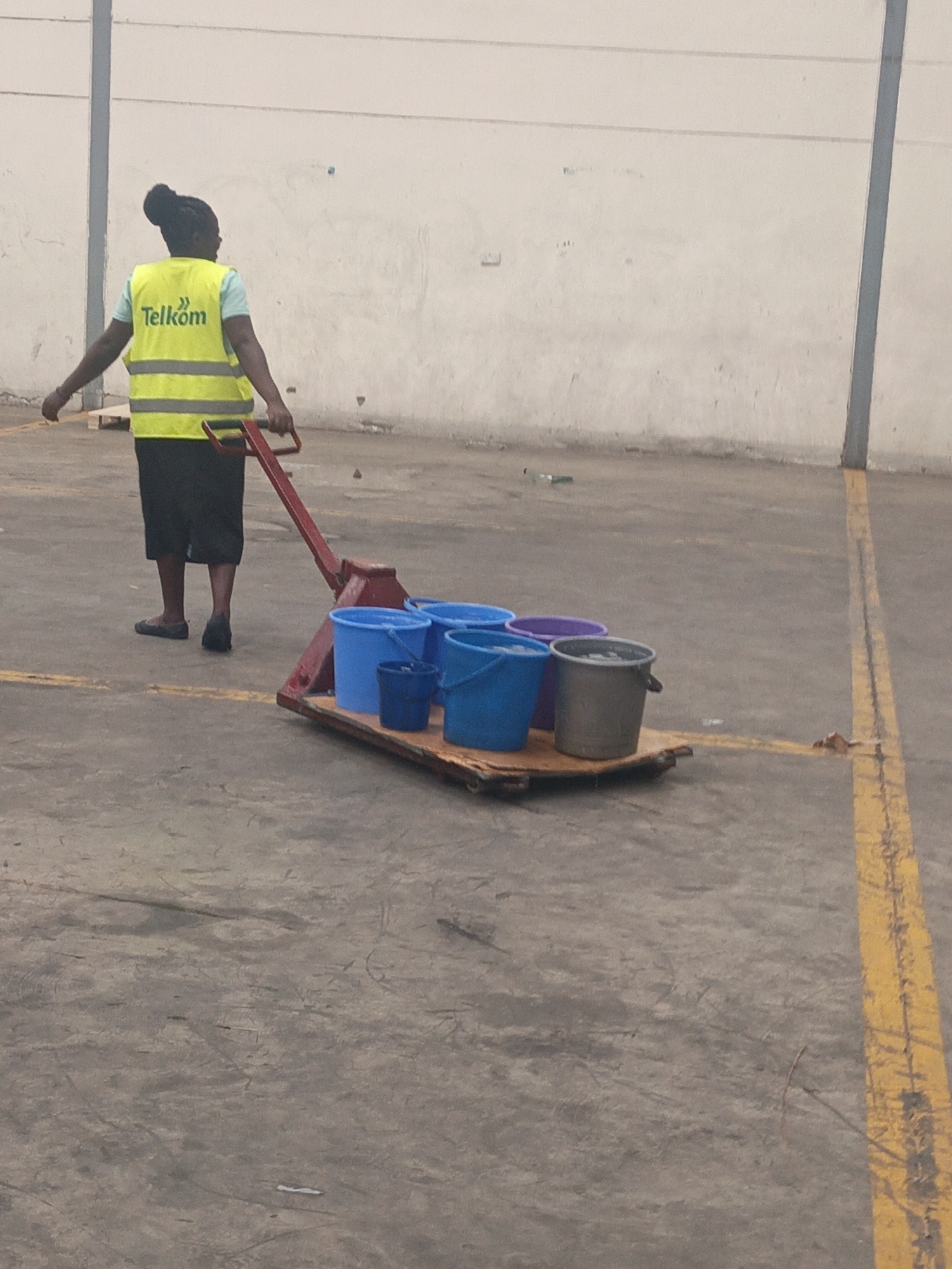 An individual wearing a yellow Telkom safety vest is pulling a red pallet jack loaded with multiple buckets of varying colors and sizes across a concrete floor. The background consists of a plain, off-white wall with a support beam. A yellow painted line runs along the right side of the frame. The location is Nairobi, Kenya.