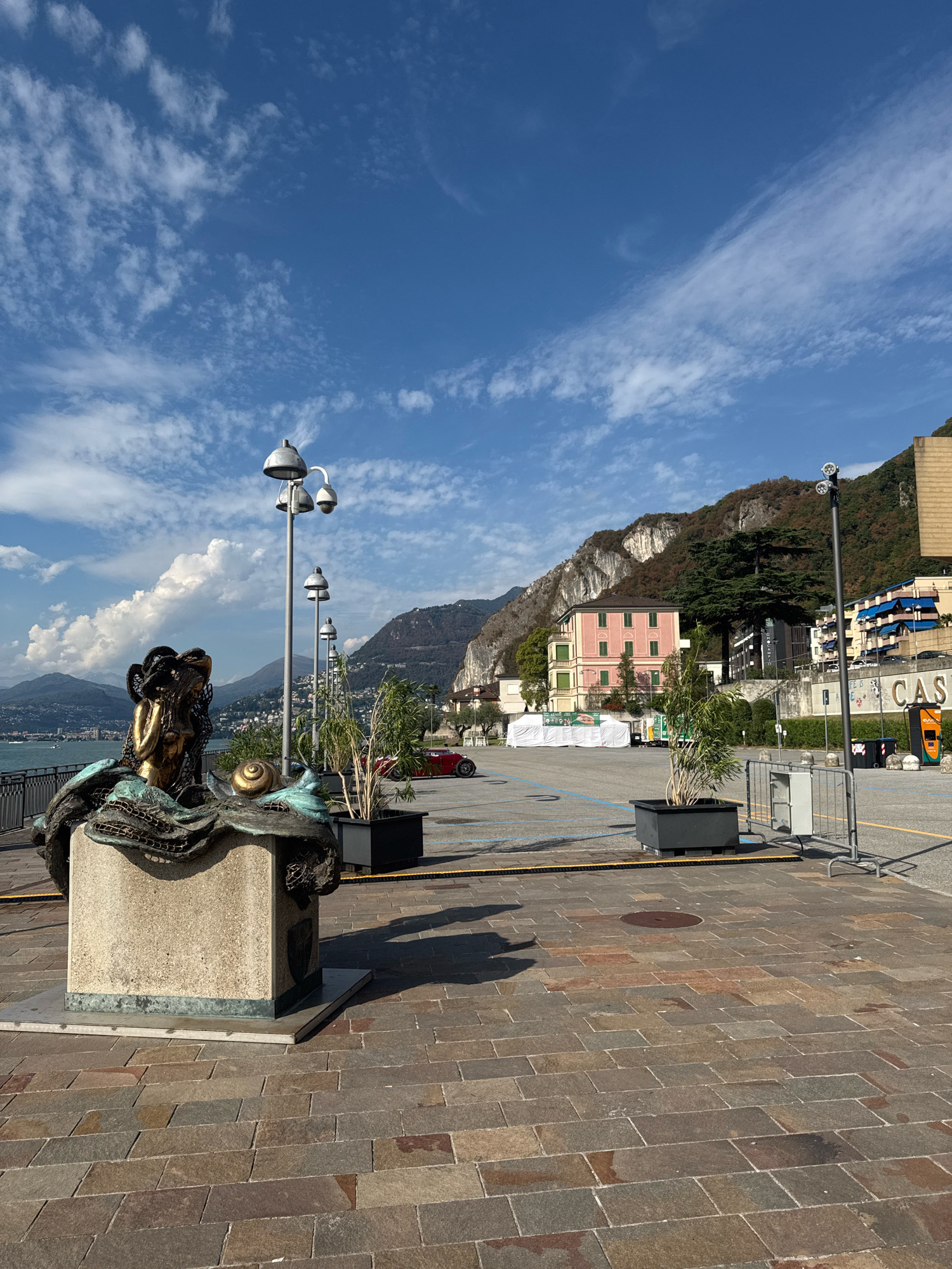 The image depicts a waterfront promenade in Campione d'Italia, Italy, with a view of Lake Lugano and the surrounding mountains. In the foreground, a bronze sculpture of a mermaid-like figure with a golden head sits on a stone pedestal. Adjacent to the sculpture is a snail. Several lampposts with multiple lights line the promenade, leading towards a parking area.
Behind the promenade, there is a parking lot with blue lines marking spaces. A red car is visible parked in the lot. Buildings of various architectural styles are situated against the backdrop of a steep, tree-covered mountain. One building is painted pink with green shutters, while others are modern structures with balconies.
The sky is a clear blue with scattered white clouds. The promenade itself is paved with stone tiles. The overall scene suggests a sunny day in a scenic, urban waterfront setting.