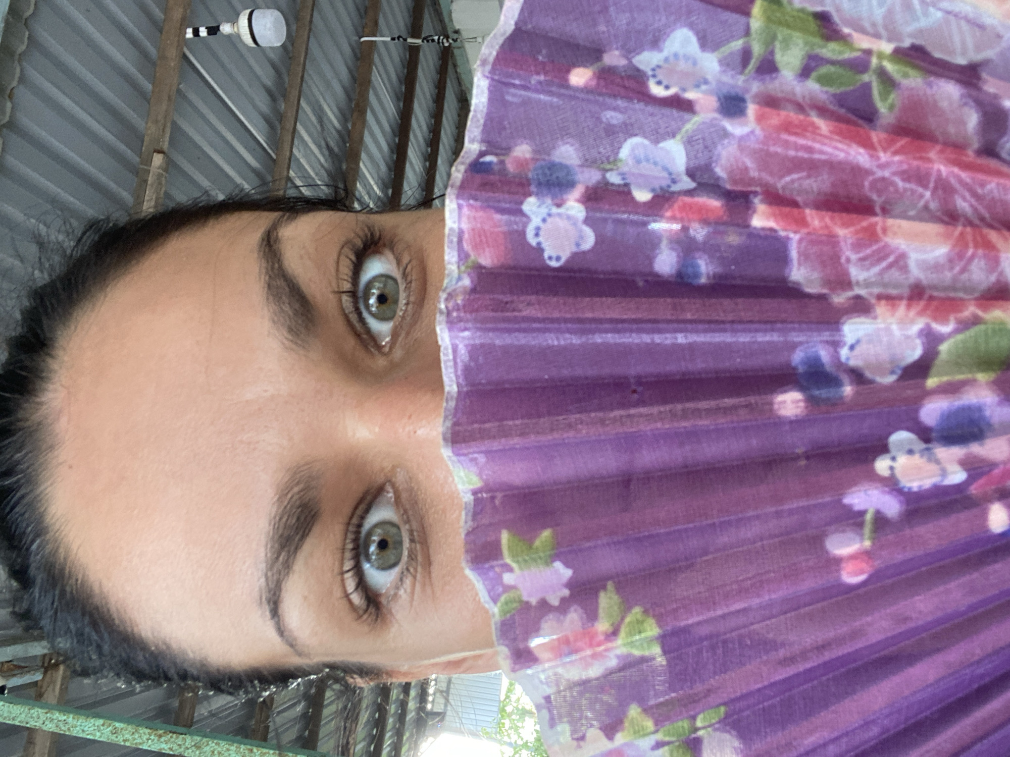 The image captures a close-up of a person's face, oriented vertically, with the upper portion of their face, primarily the eyes and forehead, visible on the left side. The individual has wide, light-colored eyes (appearing green or hazel) and prominent eyebrows, looking directly at the camera. The right side of their face is covered by a purple folding fan adorned with a floral pattern featuring light purple, pink, and white flowers with green leaves.
The background reveals a corrugated metal structure, likely a roof or wall, beneath which wooden beams or rafters are visible. In the upper left, a white light fixture with a black and white striped attachment is affixed to a beam. A green metal bar or frame runs along the bottom edge of the frame. The bright illumination suggests a daytime setting. The scene appears to be an indoor or semi-outdoor area, possibly a shed or covered space. The presence of the fan might suggest warm weather. No text is visible in the image.