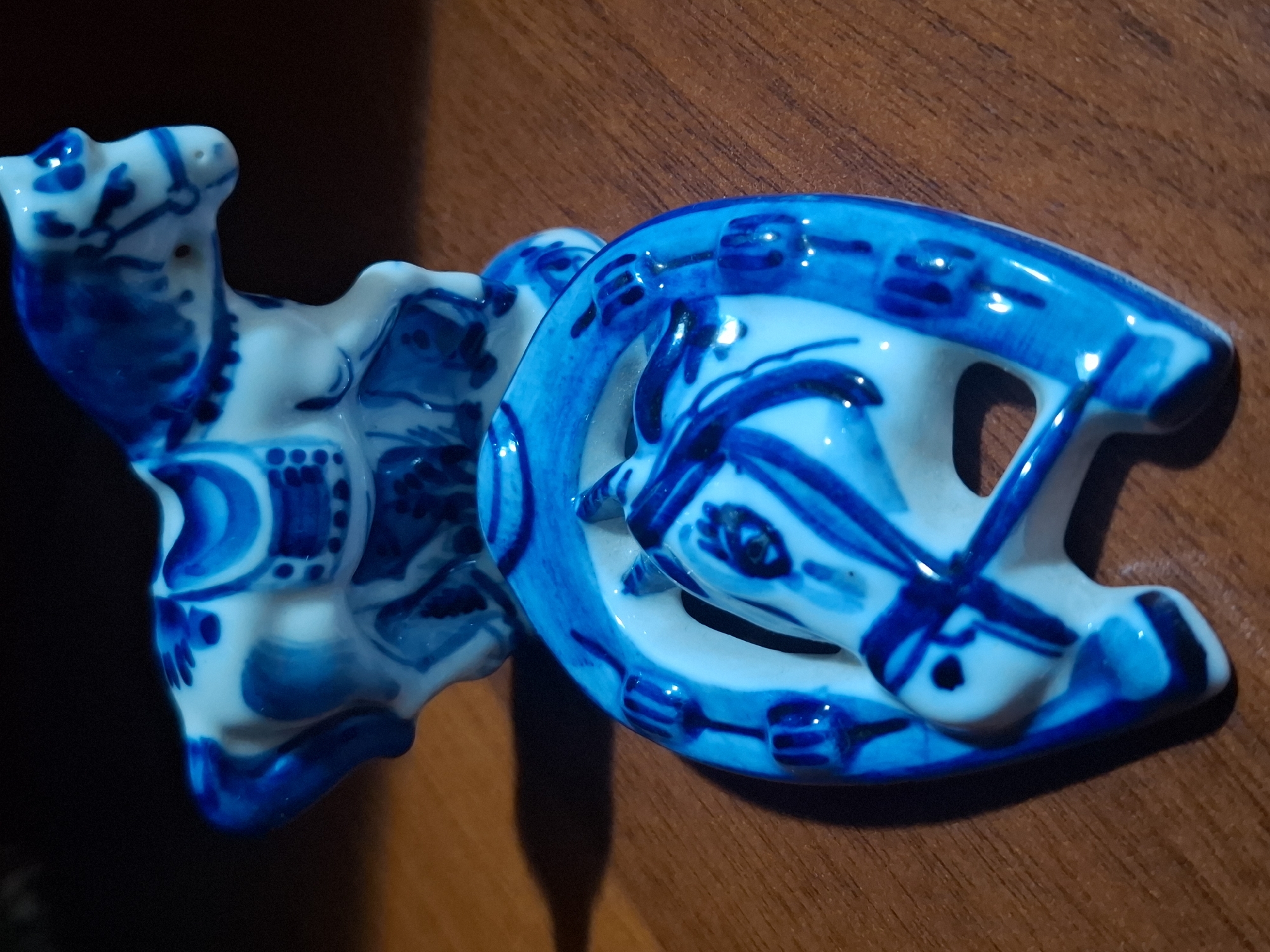 Two blue and white ceramic horse figurines are displayed on a wooden surface. One figurine, a horse with a saddle, is positioned in the background. In the foreground, a horseshoe-shaped object with a horse's head emerging from its center is placed. The figurines exhibit intricate blue floral and geometric patterns against a white background. The lighting suggests an indoor setting, with shadows cast on the wooden surface. No specific location in Almaty, Kazakhstan, can be determined from the image.