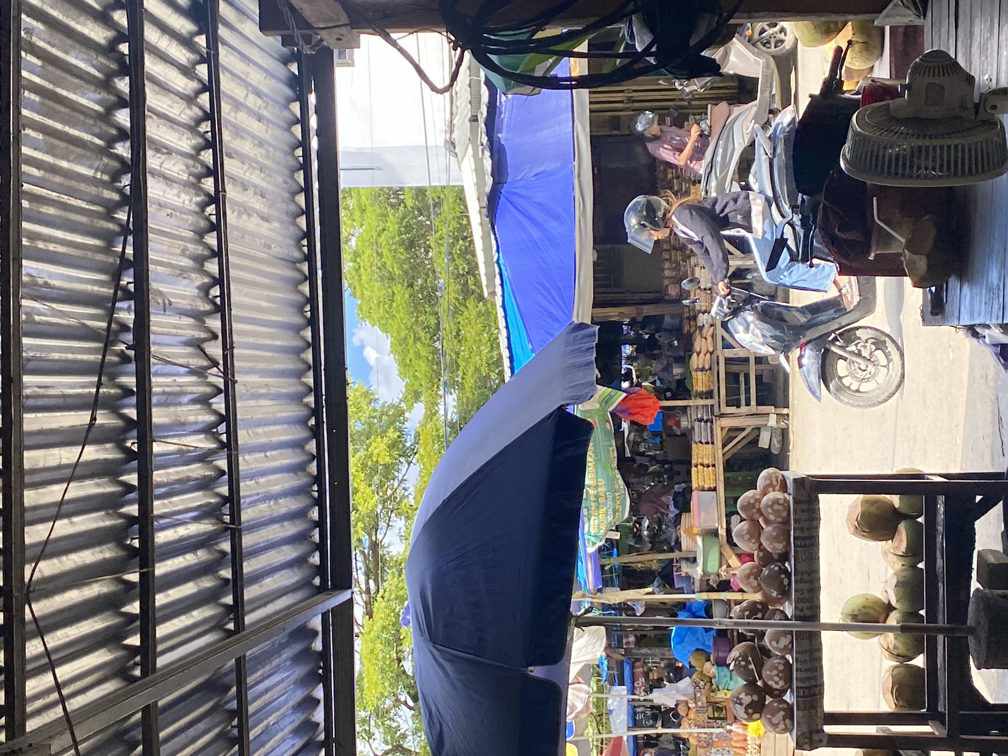 The image depicts a street market in Ta', Indonesia. A person wearing a helmet and a dark jacket rides a scooter from left to right across the frame. In the background, a blue awning covers market stalls. A large black umbrella stands to the left of the frame, partially obscuring a display of coconuts. The market appears to be outdoors, with a corrugated metal roof overhead. The overall impression is one of a busy, active marketplace on a bright, sunny day.