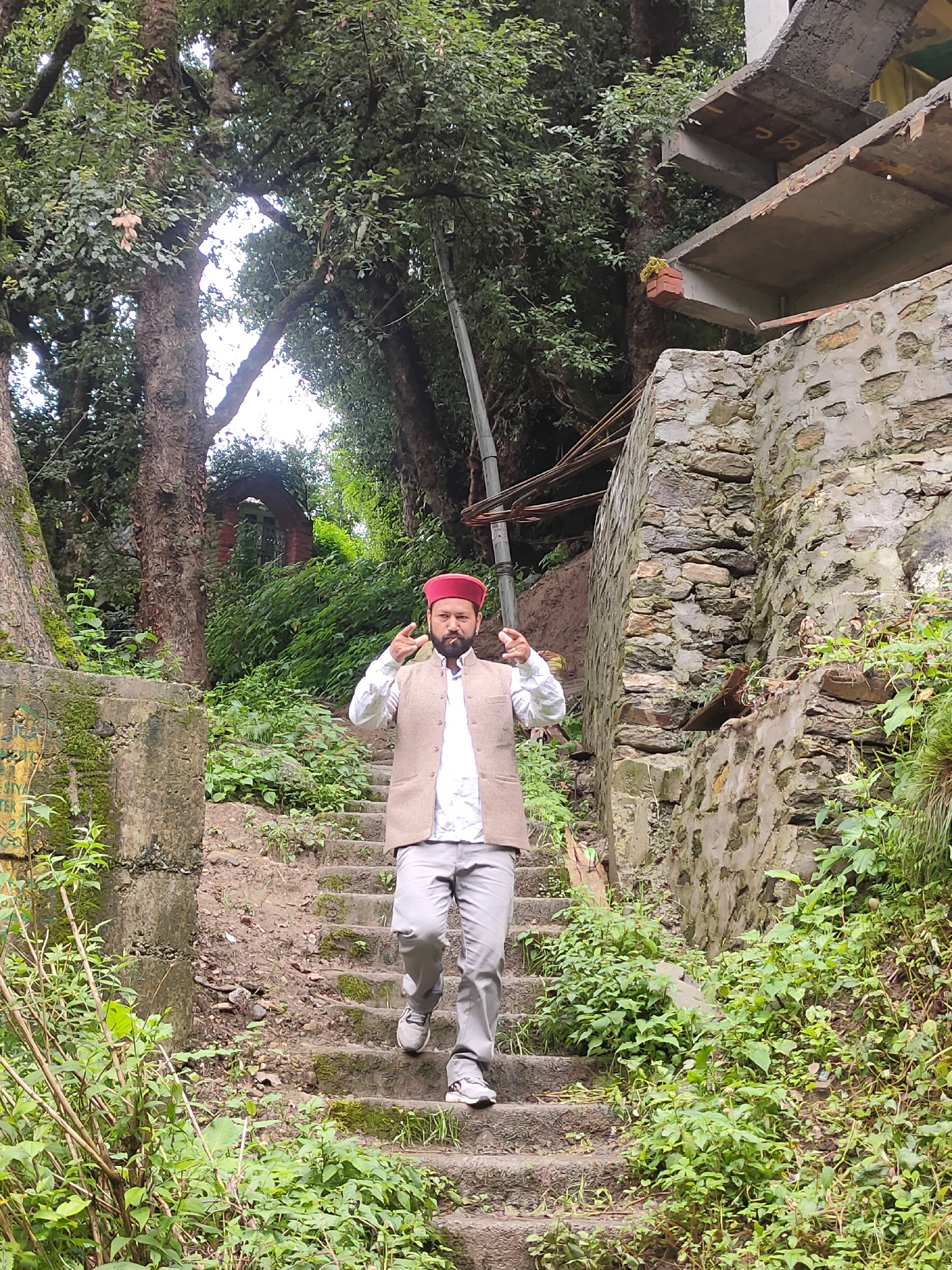 A man wearing a red cap, a white shirt, and a tan vest is walking down a stone staircase in Siyal, India. He is gesturing with his right hand while holding a cigarette in his mouth. The staircase is surrounded by lush green vegetation and trees. To the right of the staircase is a stone wall and the remains of a building.