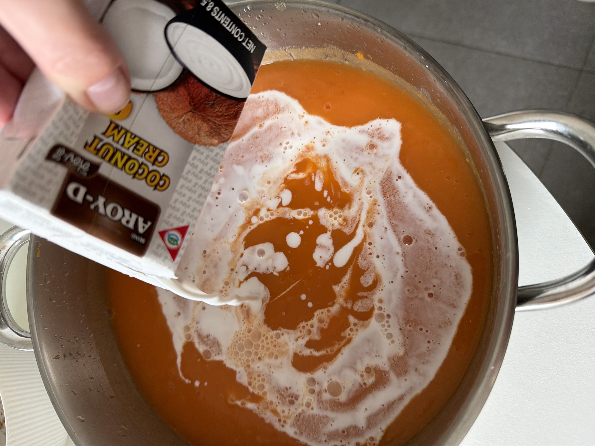 An overhead view captures an indoor cooking scene. A fair-skinned human hand, positioned in the upper right, holds a white "AROY-D Coconut Cream" carton. A steady stream of opaque white liquid is being poured from the carton into a large, round, stainless steel pot that dominates the central and lower frame. The pot contains a vibrant orange, viscous liquid, likely a soup or puree. The interaction of the pouring white liquid with the orange liquid creates distinct swirling patterns and foamy white bubbles on the surface. The carton's label clearly displays "AROY-D", "COCONUT CREAM" in yellow lettering, an image of a coconut, and "NET CONTENTS 8.5". The pot features a metallic rim and a visible curved handle on its left side. The environment consists of a light-colored, possibly white, countertop surface beneath the pot, with a portion of a white, ribbed container visible in the upper left. The specific location within Tel-Aviv, Israel, cannot be determined from the visual information presented.