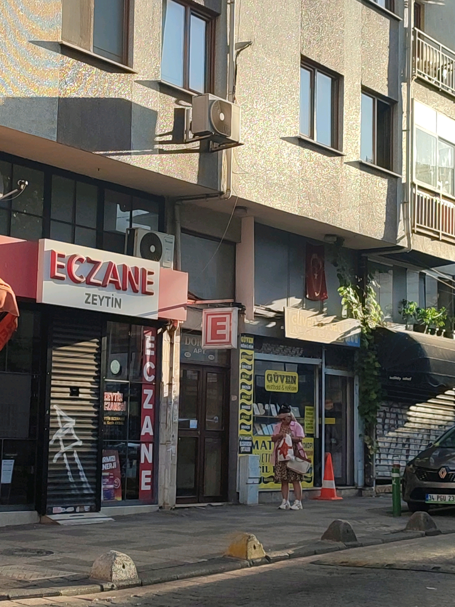 The image depicts a street scene in Kadıköy, Turkey. In the foreground, a sidewalk is visible with a few bollards. On the right, the front of a dark-colored car with the license plate "34 PGU 230" is parked. A woman in a pink top and a leopard-print dress is standing on the sidewalk, holding a shopping bag and looking at her phone. She is near an orange traffic cone. Behind her, several storefronts are visible. One prominent sign reads "ECZANE ZEYTİN," indicating a pharmacy. Another shop has a sign that says "GÜVEN matbaa & reklam." The buildings appear to be made of concrete and have windows and balconies. An air conditioning unit is mounted on one of the building walls. The scene is illuminated by sunlight, casting shadows on the pavement and buildings.