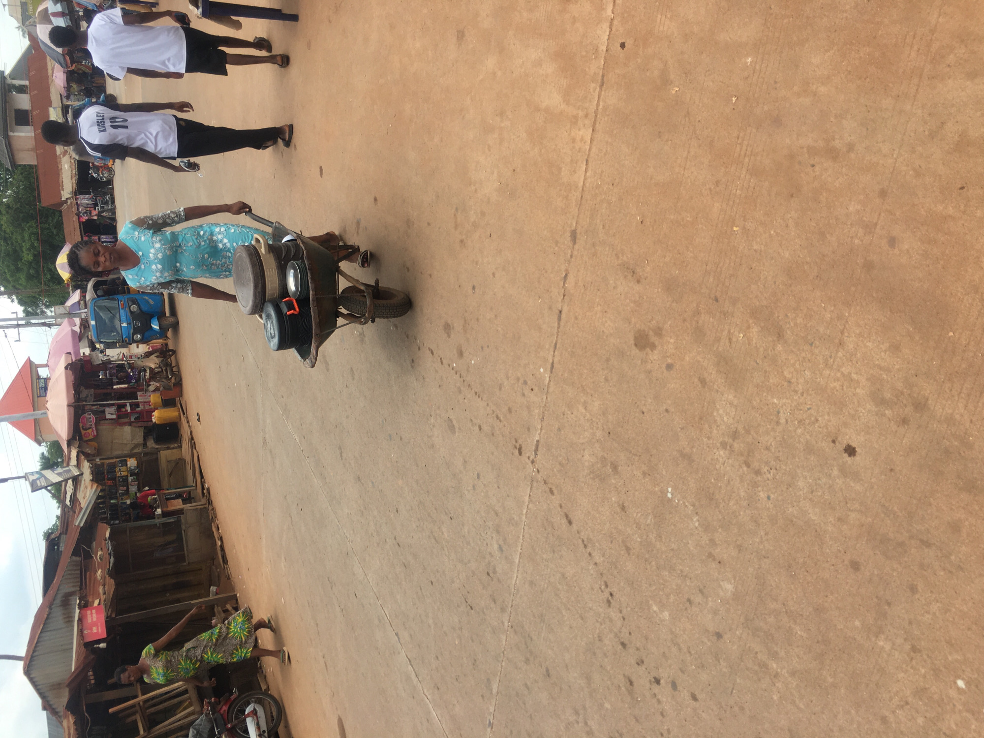 The image depicts a street scene in Enyibichiri, Nigeria. A woman wearing a blue dress walks along a concrete road, pushing a wheelbarrow loaded with metal pots and pans. Two young men wearing white shirts walk in the same direction, parallel to the woman. In the background, there is a blue "Keke Napep" (tricycle taxi) parked near stalls and shops. One stall has a variety of shoes displayed. A second woman in a green patterned dress is standing next to a store. A motorcycle is parked next to the store.