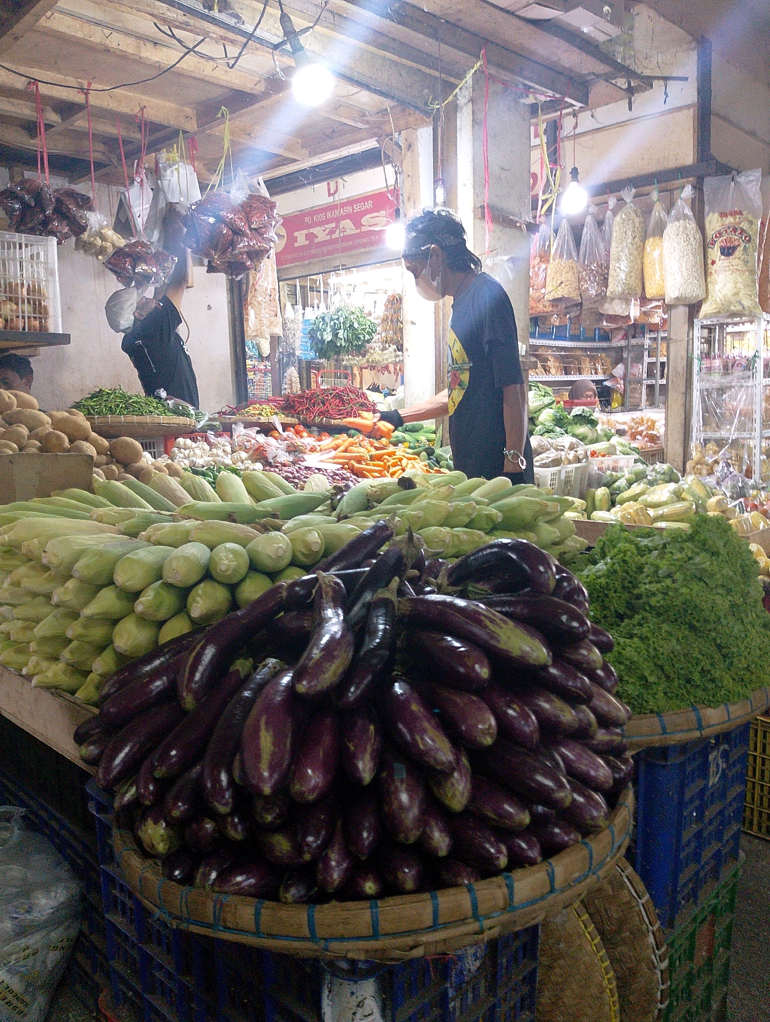 The image depicts a vibrant market stall in Lembang, Indonesia, filled with a colorful array of fresh produce. In the foreground, a large pile of deep purple eggplants is neatly arranged in a woven basket, some of them appearing slightly glossy. To the left and behind the eggplants, a substantial mound of corn on the cob, still in its husks, dominates the scene. Further back, other vegetables like green beans, carrots, and chili peppers are visible, along with sacks of potatoes and heads of garlic.
A person wearing a dark shirt and a light-colored face mask is partially visible on the right side of the frame, seemingly interacting with the produce or attending to customers. The market stall is illuminated by bright overhead lights, casting a warm glow on the scene. The background reveals more shelves stocked with various goods, including bags of snacks and spices, and the general ambiance suggests a bustling local marketplace. The overall impression is one of abundance and lively commerce.