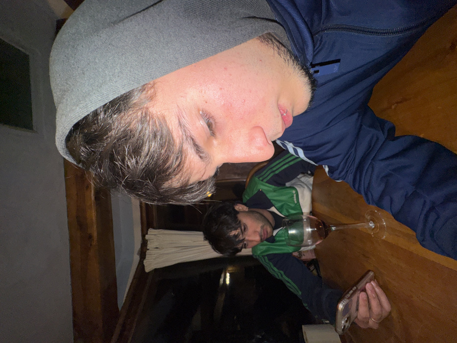 Two men are captured in a dimly lit interior. One, wearing a grey beanie and a dark blue tracksuit, appears closer to the camera. Below him, a second man is visible in the image, reflected in the glass of a table. He's wearing a green and blue tracksuit, holding a cell phone in his hand. A wine glass is situated between them. The image is captured at an awkward angle, with the subjects seemingly tilted. A window with drawn white curtains is visible in the background.