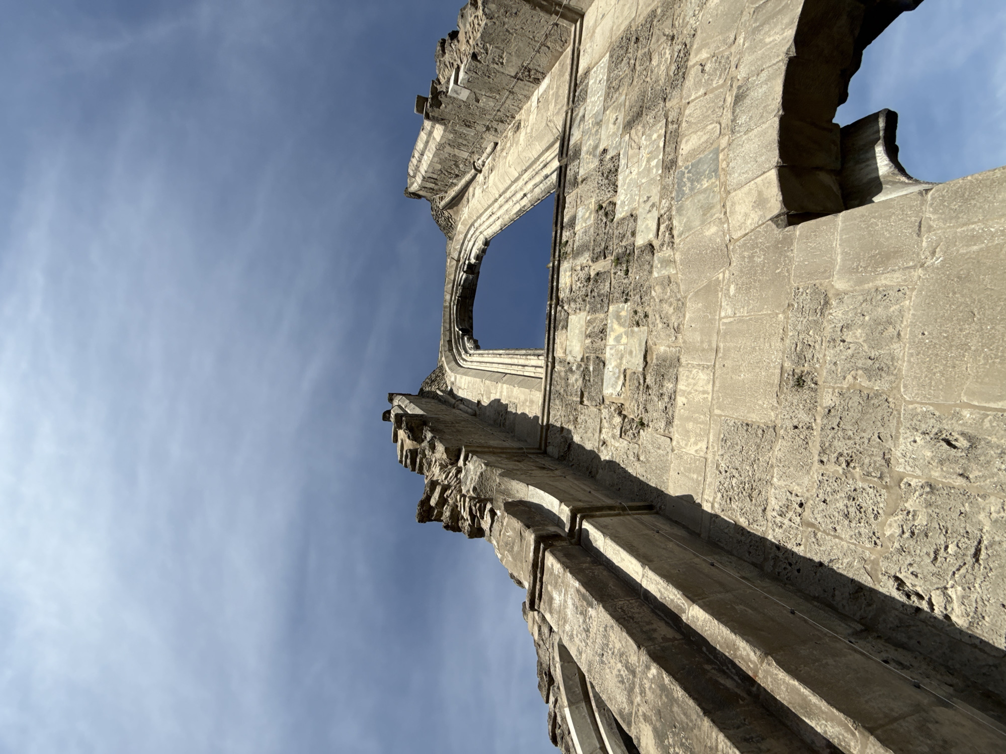This is a low-angle, full outdoor shot of the dilapidated ruins of the Walkenried Abbey, located in Walkenried, Germany. The sky above is a bright blue with scattered wispy clouds. The focus is on the weathered stone architecture of the abbey. The building exhibits signs of significant decay, with crumbling edges and missing sections of stone. An arched window opening is clearly visible, framing the sky. The stonework appears to be large, roughly hewn blocks, some of which are discolored and textured with age and exposure to the elements. Shadows cast by the sun highlight the architectural details and the uneven surfaces of the stone. No people are present in the frame.