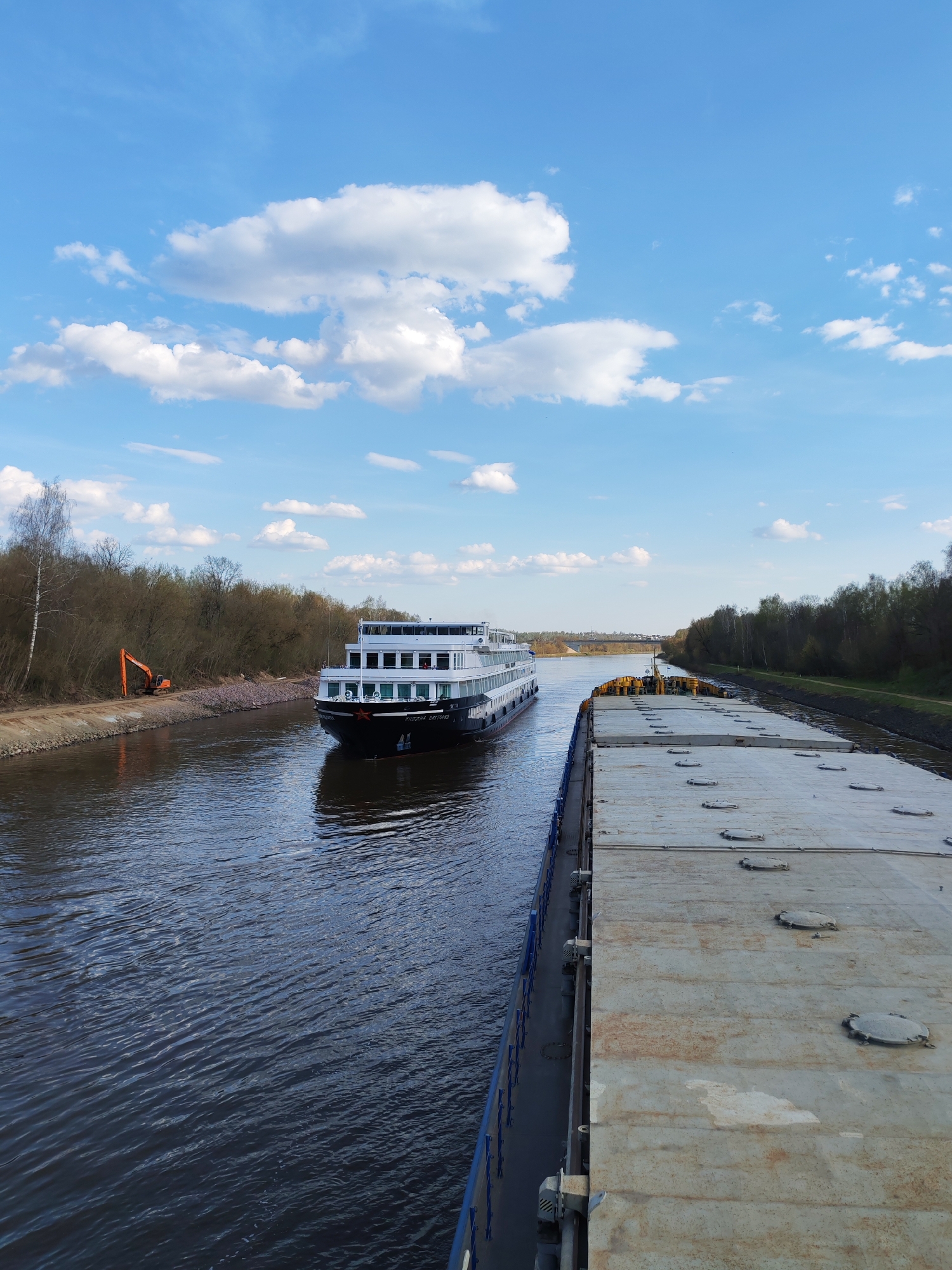The image shows a canal in Varvarino, Russia. A multi-decked river cruise ship passes a large barge loaded with concrete panels. On the left bank, an orange excavator sits along the edge of the trees. The sky is blue with scattered white clouds.