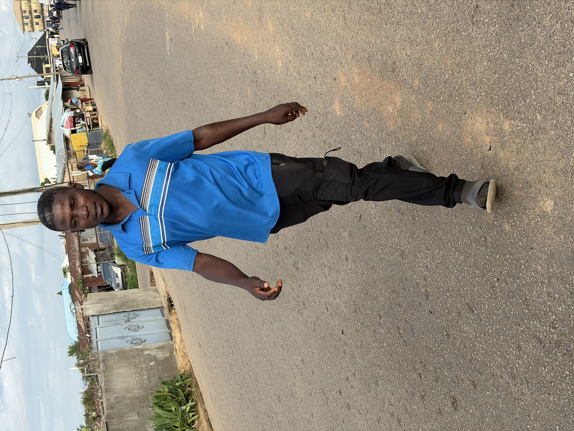 An outdoor scene in Jos, Nigeria, depicts a Black male in his late 20s to early 40s walking forward on an asphalt road. He wears a blue polo shirt with horizontal white and light blue stripes across the chest, black cargo trousers with a zippered pocket, and grey sandals with white soles. His expression is neutral, and he looks towards the camera.
To his left, a concrete wall topped with barbed wire encloses a property, featuring a light blue metal gate with decorative patterns. Green foliage is visible behind the wall, and a light blue corrugated metal roof of a building is discernible further back. A silver SUV is partially visible behind the gate. Overhead, utility lines and poles traverse the scene against an overcast sky with patches of blue.
To his right, further down the road, a dark-colored hatchback car (possibly a Renault Clio) drives away. The road is lined with various structures, including makeshift market stalls with corrugated metal roofs and colorful umbrellas, where several individuals are visible. Other buildings of varying architecture extend into the distance, indicating a busy urban or semi-urban environment. The asphalt road surface shows wear and scattered aggregate. The scene is illuminated by natural daylight.