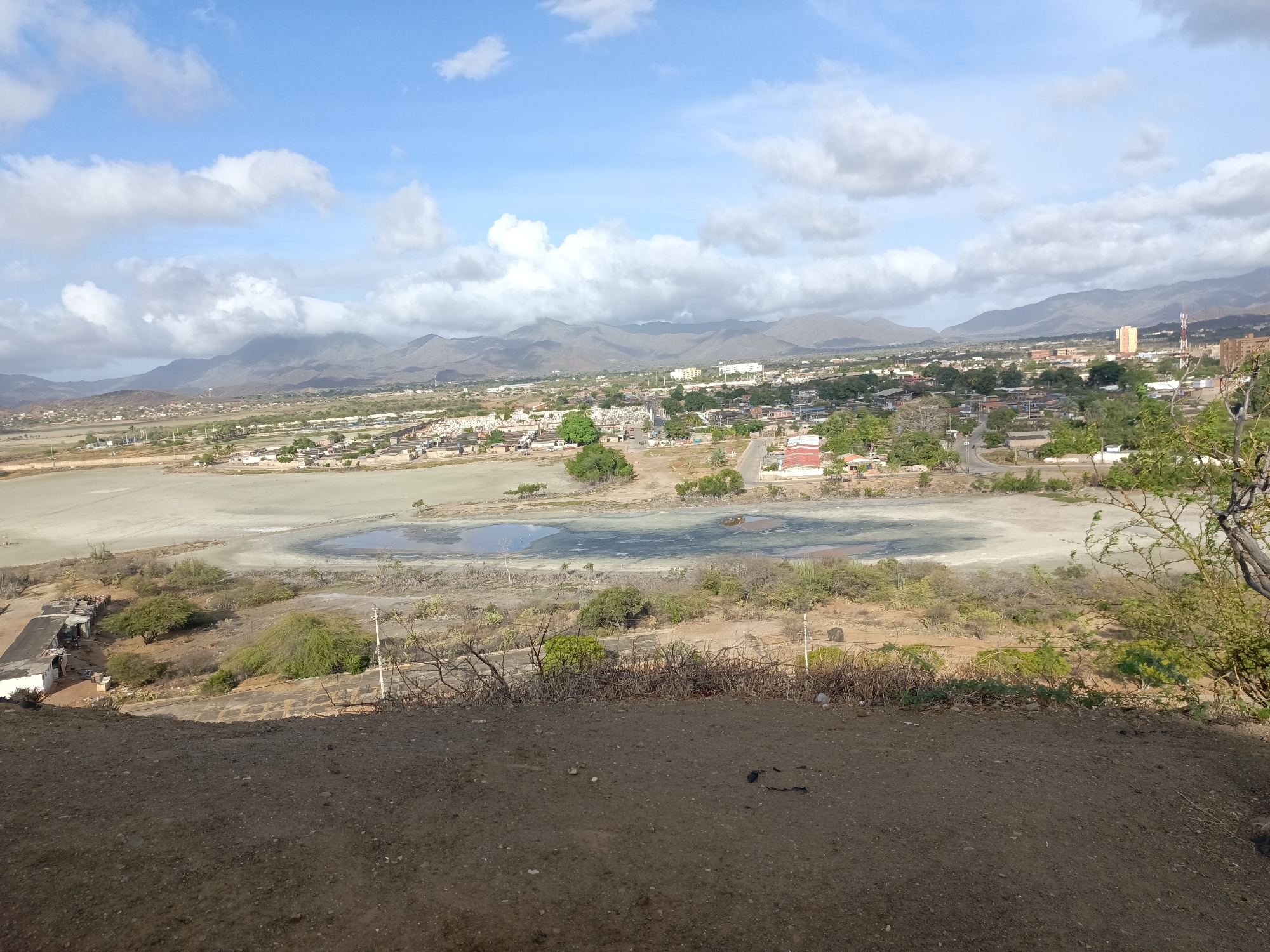 The image depicts a panoramic view of Juan Griego, Venezuela, from an elevated vantage point. The foreground is dominated by a brown, earthy slope. Beyond this, a large, shallow body of water extends into the midground, appearing somewhat dried up with varying shades of gray and patches of stagnant water.
The city of Juan Griego is visible in the background, characterized by a mix of residential and commercial buildings. Patches of trees and green spaces are interspersed among the structures. In the distance, a mountain range forms a backdrop under a partly cloudy sky. A prominent tower stands out among the buildings in the city.