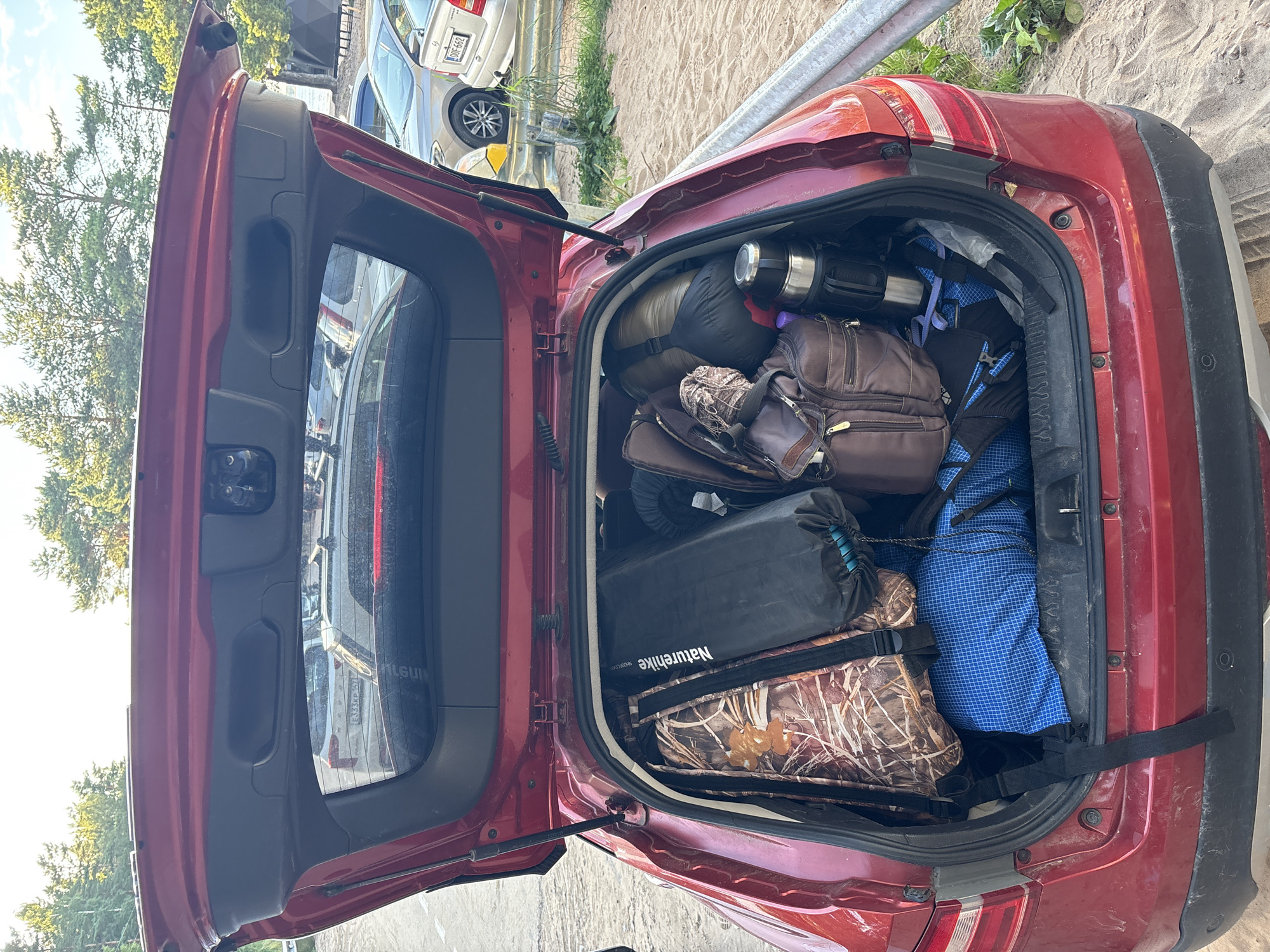 The image shows the open trunk of a red car, packed with camping gear. The trunk contains several items, including a camouflage-patterned bag, a black sleeping bag, a blue checkered duffel bag, a brown backpack, and a silver thermos. The car is parked on a sandy surface, with trees and other cars visible in the background. The scene appears to be in a natural outdoor setting, possibly a campsite or a parking area near a park. The lighting suggests it is daytime, with sunlight filtering through the trees. There are no people visible in the image. The location is identified as Kokkorevo, Russia.