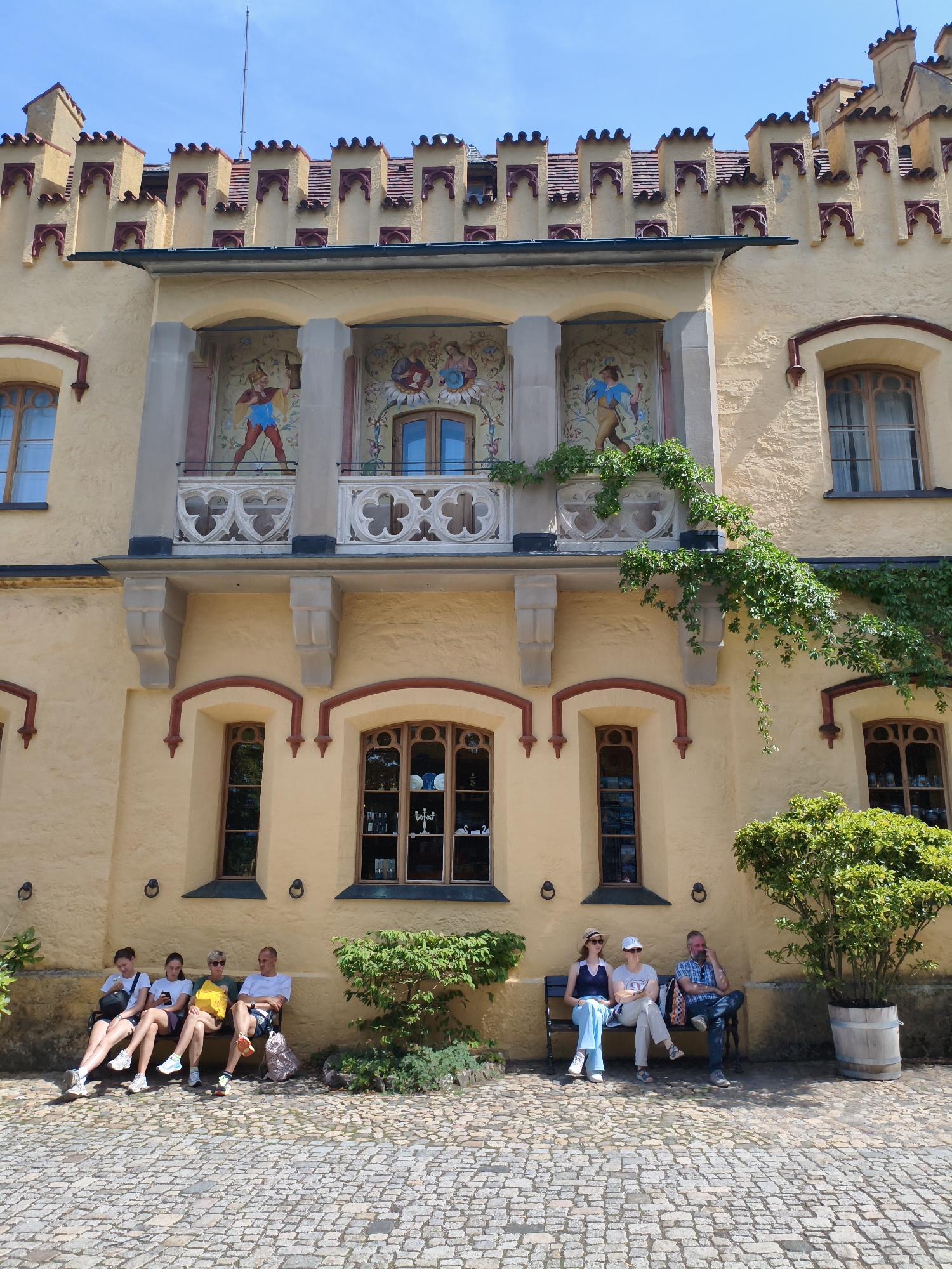 The image shows the exterior of a yellow castle with red-trimmed battlements against a clear blue sky. In the foreground, a group of seven people, varying in age and gender, are seated on two benches on a cobblestone patio. Some are looking at their phones, while others are conversing or resting. The castle features ornate windows and decorative murals on an upper balcony, with lush green vines growing on its facade. The scene appears to be a tourist location, likely during the daytime given the bright sunlight and shadows. The location is identified as Schwangau, Germany, a town known for its fairy-tale castles.