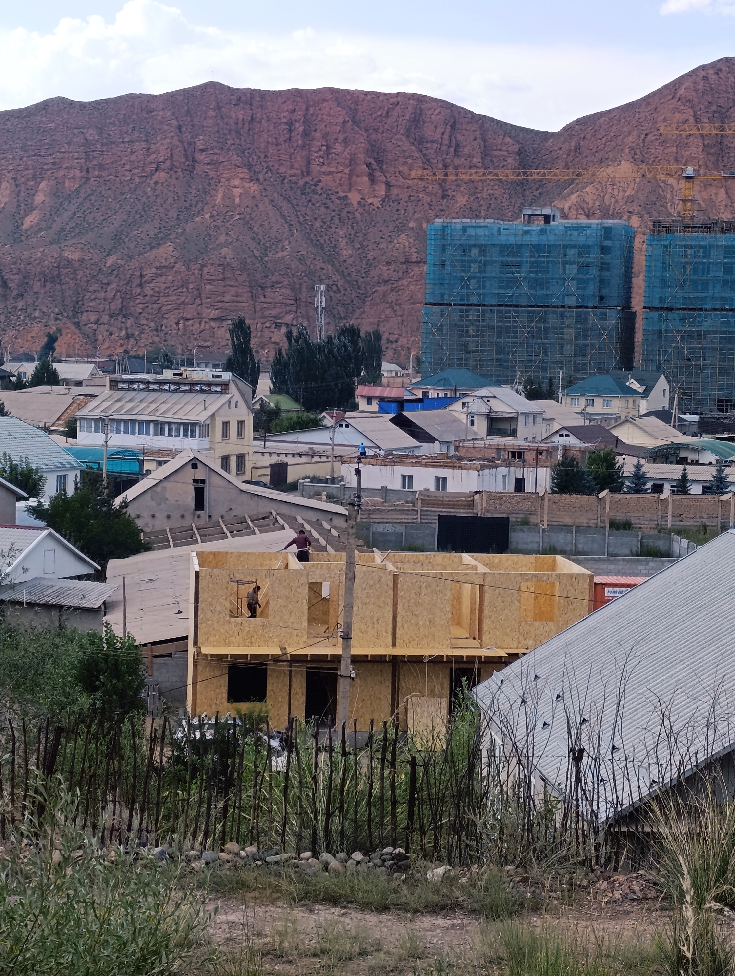 In the city of Naryn, Kyrgyzstan, this image captures a construction site with a partially built wooden structure in the foreground, featuring two individuals working. The surrounding environment includes a fence made of vertical wooden poles and dry vegetation. In the midground, a collection of residential houses with diverse roof designs are visible. Further in the background, two tall buildings are under construction, shrouded in blue netting, with yellow cranes positioned nearby. Towering over the scene are arid, reddish-brown mountains, characteristic of the landscape surrounding Naryn. The sky is partly cloudy.