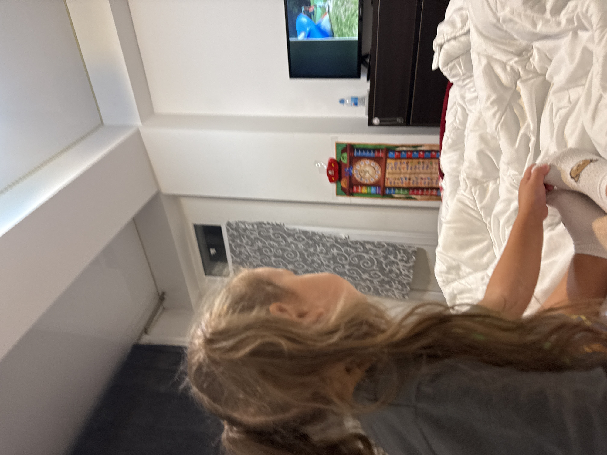 The image shows a young girl with blonde hair in a ponytail, facing away from the camera and looking at a television screen. She is wearing a grey t-shirt and white socks. The girl is sitting on a bed covered with white bedding. A dark dresser with a television on top is visible in the background. A decorative clock is hanging on the wall to the left of the dresser. The environment appears to be a bedroom. The location is not specified in the image.