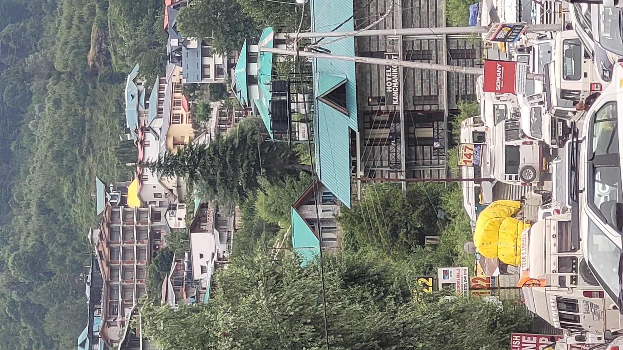 This image depicts a street scene in Vashisht, India, characterized by a dense collection of buildings and vehicles. The architecture features a mix of styles, with several buildings topped by turquoise-colored, sloped roofs, common in hilly regions. Some buildings appear to be hotels or guesthouses, as indicated by the sign for "HOTEL KANCHANIKO" and other visible signage related to "TOUR & TRAVEL."
The street is lined with numerous white vans and cars, suggesting a busy thoroughfare, possibly a tourist area with shuttle services. A bright yellow, voluminous object, possibly luggage or a tarpaulin, is prominently placed near some of the vehicles. The background shows a steep, densely forested hillside, indicative of a mountainous or hilly terrain.
Visible text on signs includes hotel names, service advertisements like "SOMANY," and route numbers such as "147." There are also signs with numbers like "22." The overall impression is a bustling, commercial street in a tourist destination nestled within a natural landscape. The lighting suggests it could be daytime, with overcast conditions contributing to a soft illumination. The scene captures the vibrancy and activity of a popular Indian tourist spot.