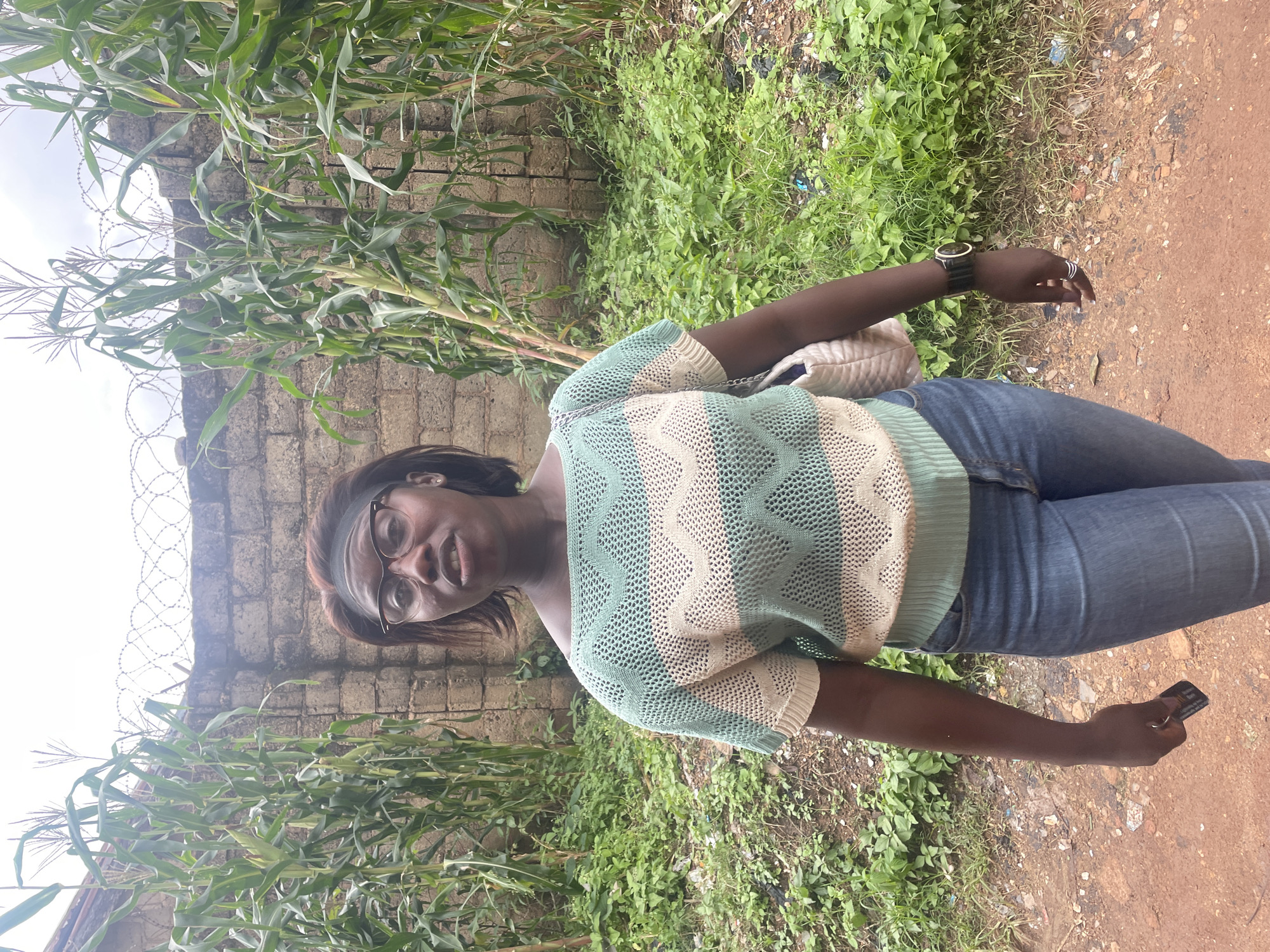 In Jos, Nigeria, a woman with dark skin and short, dark hair stands in front of a brick wall topped with barbed wire. She is wearing glasses, a striped green and white off-the-shoulder sweater, and blue jeans. A cream-colored purse hangs from her left shoulder. In her right hand, she holds a black object with a silver keychain attached. The background features tall corn stalks and lush green vegetation. The ground is a mix of reddish-brown dirt and patches of grass with scattered debris.