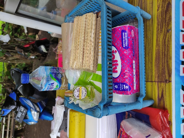The scene is a street food vendor stall in Kecamatan Lawang, Indonesia (latitude: -7.834, longitude: 112.693). The vendor is selling food items from a blue metal shelf. The shelf is positioned on a wooden table. The shelf contains a box of tissue paper, a stack of wooden skewers, and a bottle of clear liquid. A blue scooter is visible in the background, partially obscured by vegetation. There is also a partially obscured yellow object on the table in the foreground. The environment is outdoors, likely on a street or sidewalk.