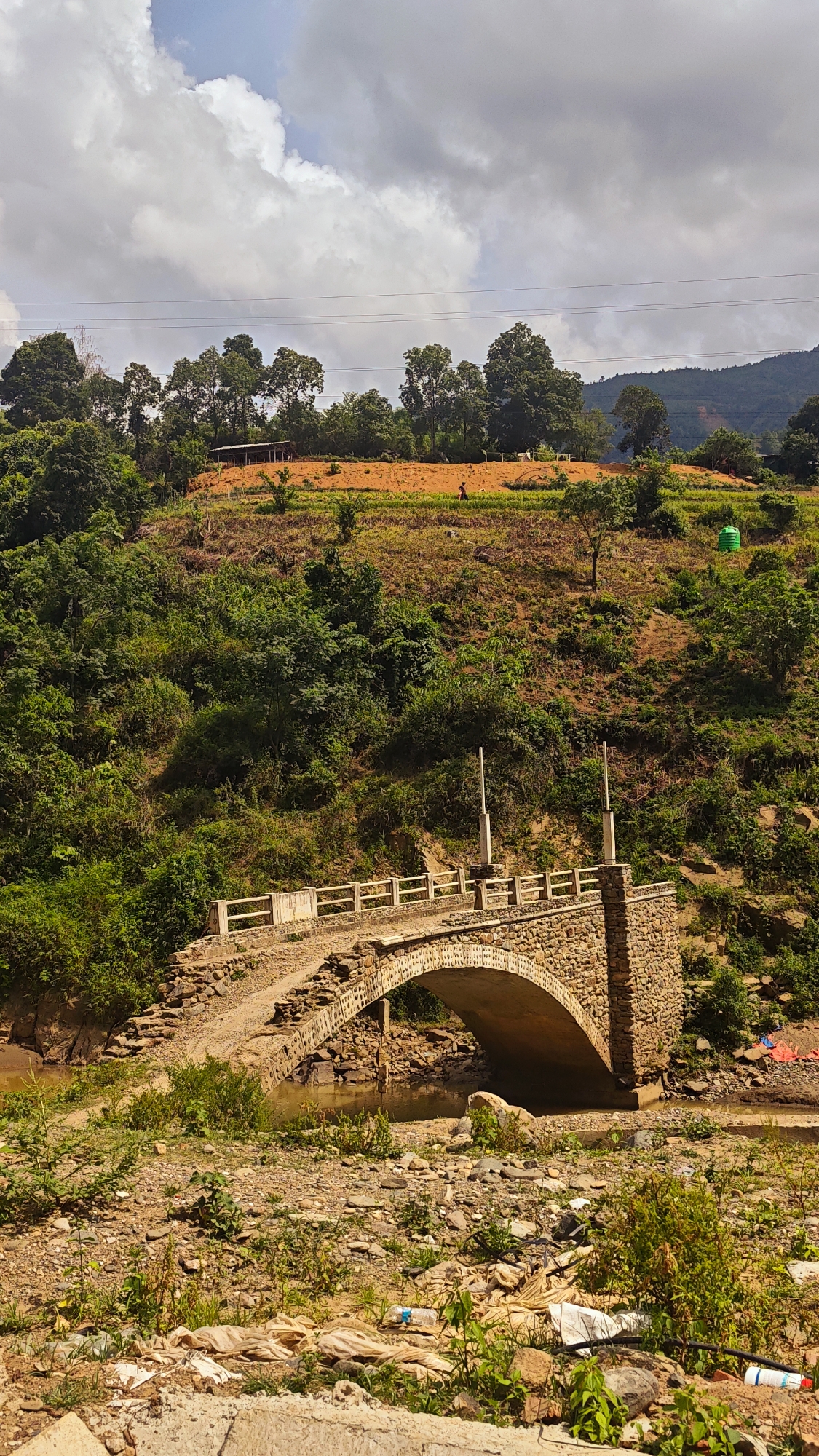 This vertical shot captures a rural landscape in Khao Mang, Vietnam, featuring a stone arch bridge in the foreground and verdant hills under a cloudy sky.
The central subject is an old-style stone arch bridge, constructed from rough-hewn stones, spanning a shallow, brownish river or stream bed. The bridge has a slightly raised, unpaved path and features simple white or light-colored concrete railings. Two vertical posts stand on the bridge deck, possibly remnants of old lampposts. The riverbed below the bridge is rocky and appears to have low water levels, with some visible debris and litter, including plastic bottles and bags, scattered in the immediate foreground.
Beyond the bridge, steep hillsides are covered in dense green foliage, ranging from shrubs to mature trees. Higher up, patches of land are cleared and tilled, revealing reddish-brown soil, suggesting agricultural activity. On one of these tilled sections, a small human figure, likely a farmer, is visible, possibly tending to crops or walking. Further up the hillside, a small, rustic shelter with a dark roof is nestled among the trees, and a bright green water tank or barrel is also visible. Power lines stretch horizontally across the upper middle ground, supported by poles partially obscured by trees. The sky above is mostly overcast with large white and grey clouds, indicating a partly cloudy day, likely late morning or afternoon given the bright natural light.
The scene depicts a blend of traditional rural infrastructure and natural beauty, with a notable presence of modern-day litter in the immediate foreground. No visible text is present in the image.