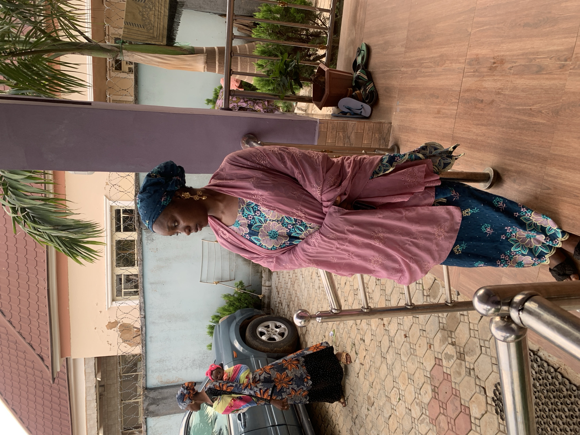 This image captures an outdoor residential scene in Jos, Nigeria, during daylight hours. In the foreground, a woman of African descent stands in profile, facing left. She wears a dark teal dress with a floral pattern, a light pink embroidered shawl draped over her shoulders, and a matching teal head wrap. Gold dangle earrings adorn her ears. She leans against a prominent purple structural pillar, with a silver metal railing extending in front of her. The ground beneath her is covered with light brown wood-grain effect tiles.
In the mid-ground, another woman of African descent walks towards the left, carrying an infant secured in a colorful fabric wrap on her back. The infant's head, wearing a pink hat, is visible above her left shoulder. This woman wears a dark-colored dress with an orange floral pattern, a head wrap, and sandals. Behind her, a dark grey SUV is parked on a patterned concrete paver driveway, which features hexagonal and octagonal interlocking blocks in shades of red, brown, and grey.
A light blue wall topped with barbed wire fencing separates the immediate foreground from a two-story house in the background. The house features a reddish-brown tiled roof and light peach-colored walls, displaying some areas of paint damage. Multiple windows are visible on the house. To the right of the main subject, several pairs of sandals and flip-flops rest on the tiled floor, and a patterned prayer mat is draped over a frond of a palm tree. Green shrubbery is present near the parked vehicle and in planters closer to the foreground.