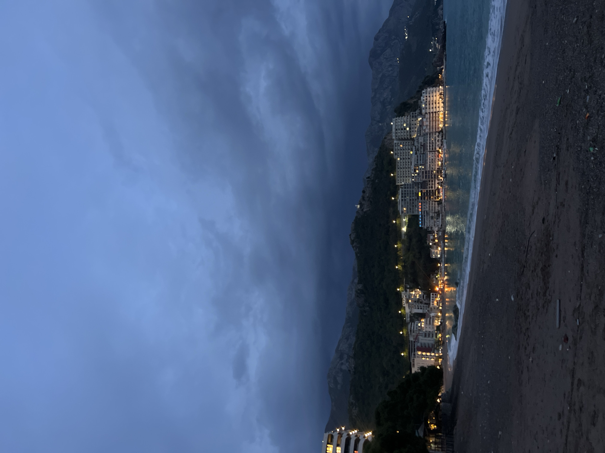 The scene is a coastal landscape at dusk or night, likely in Rafailovići, Montenegro, given the distinctive architecture clinging to the hillsides. A dark, heavy storm cloud occupies the left side of the frame, dominating the sky. A steep, verdant mountain ridge rises from the shoreline, with illuminated buildings and structures nestled into its base. These buildings display multiple lit windows, suggesting occupancy and providing the only warmth of the scene. A narrow beach with dark sand is visible in the foreground, with the shallow sea washing onto the shore. The waves are small and white-tipped. The scene is dimly lit, primarily by the artificial lights of the buildings. There are no people visible in the frame.