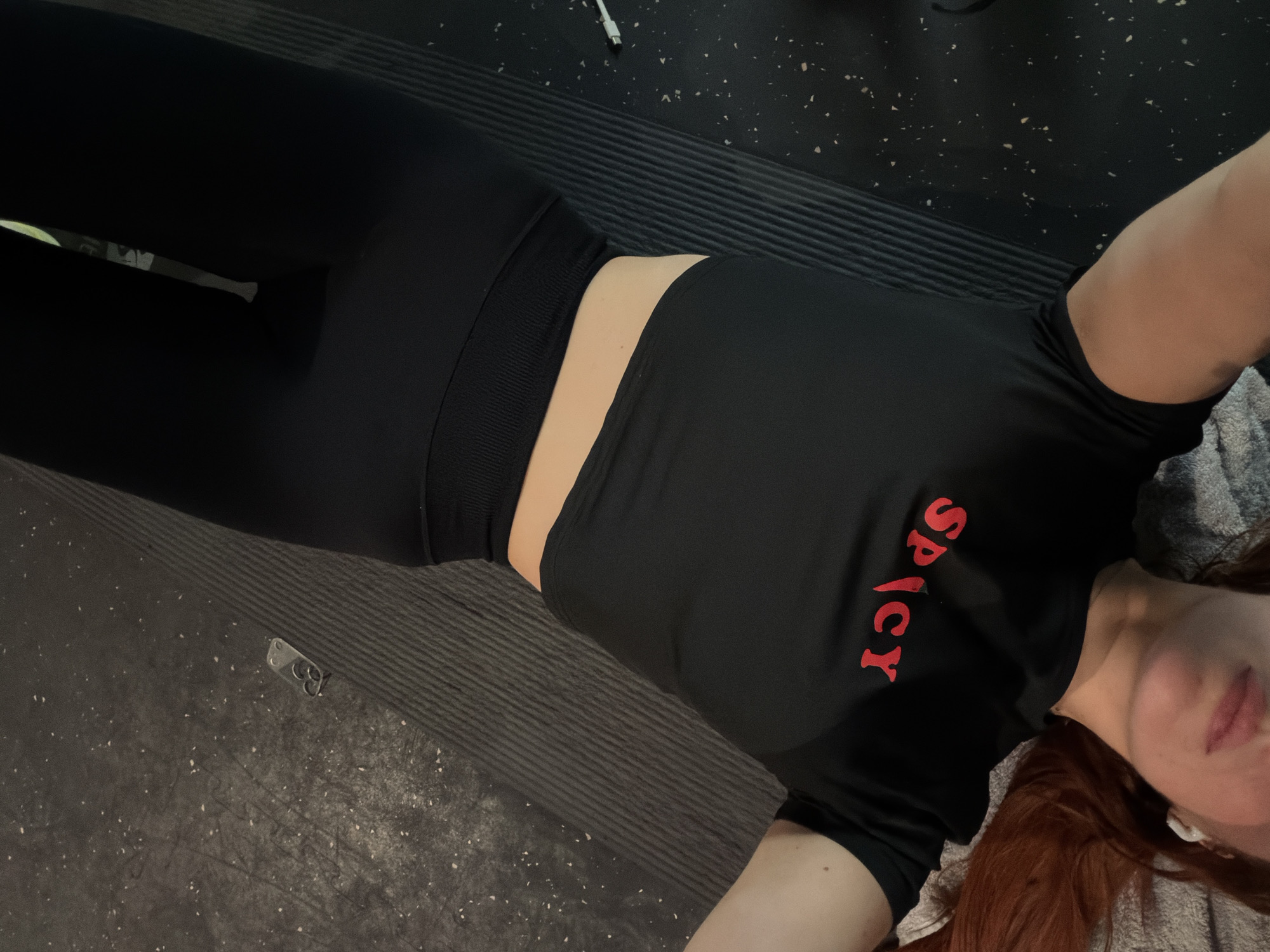 The image shows a person lying on a black textured surface. The person is wearing black leggings and a black top with the word "SPICY" in red letters. Part of their face is visible, showing red hair and a white earbud in their ear. The background includes a black floor with white specks and a grey towel. A white USB cable is also visible in the background. No information suggests it is located in Paris, France.