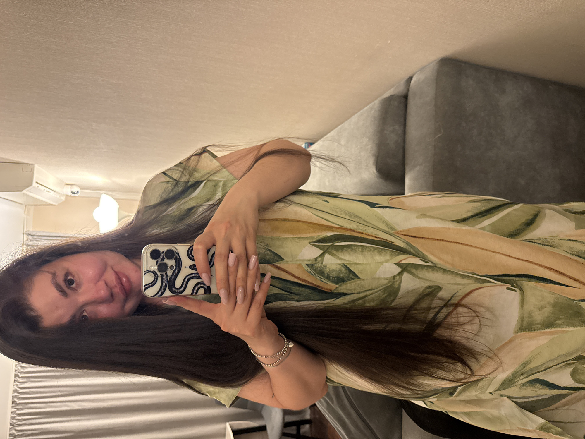 A woman takes a selfie in what appears to be an indoor living space. She is holding a smartphone with a black and white snake design case. The woman has long dark hair and is wearing a light-colored dress with a leafy pattern. A gray couch is partially visible in the background. The exact location within Votkinsk, Russia, cannot be determined from the image.