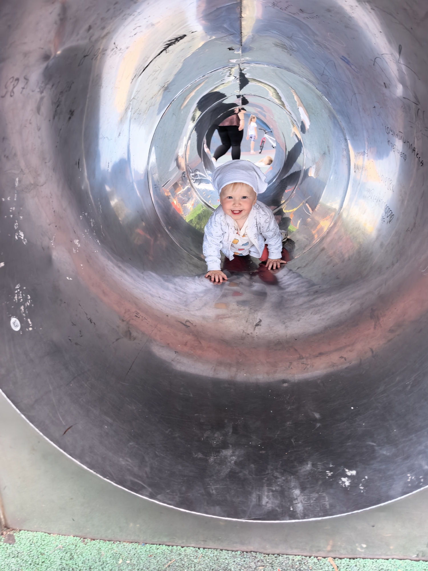 A toddler, wearing a white head covering and a light-colored patterned outfit, is crawling inside a large, silver, tubular slide. The toddler is smiling and looking directly at the camera. In the background, further down the slide, an adult is visible along with another child. The interior of the slide shows reflections of the surroundings and some graffiti or markings on the metal surface. The bottom of the slide appears to be covered with green rubber mulch. The scene suggests a playground or amusement park. There is no specific indication of the location within the city of Sterlitamak, Russia, in the image itself.