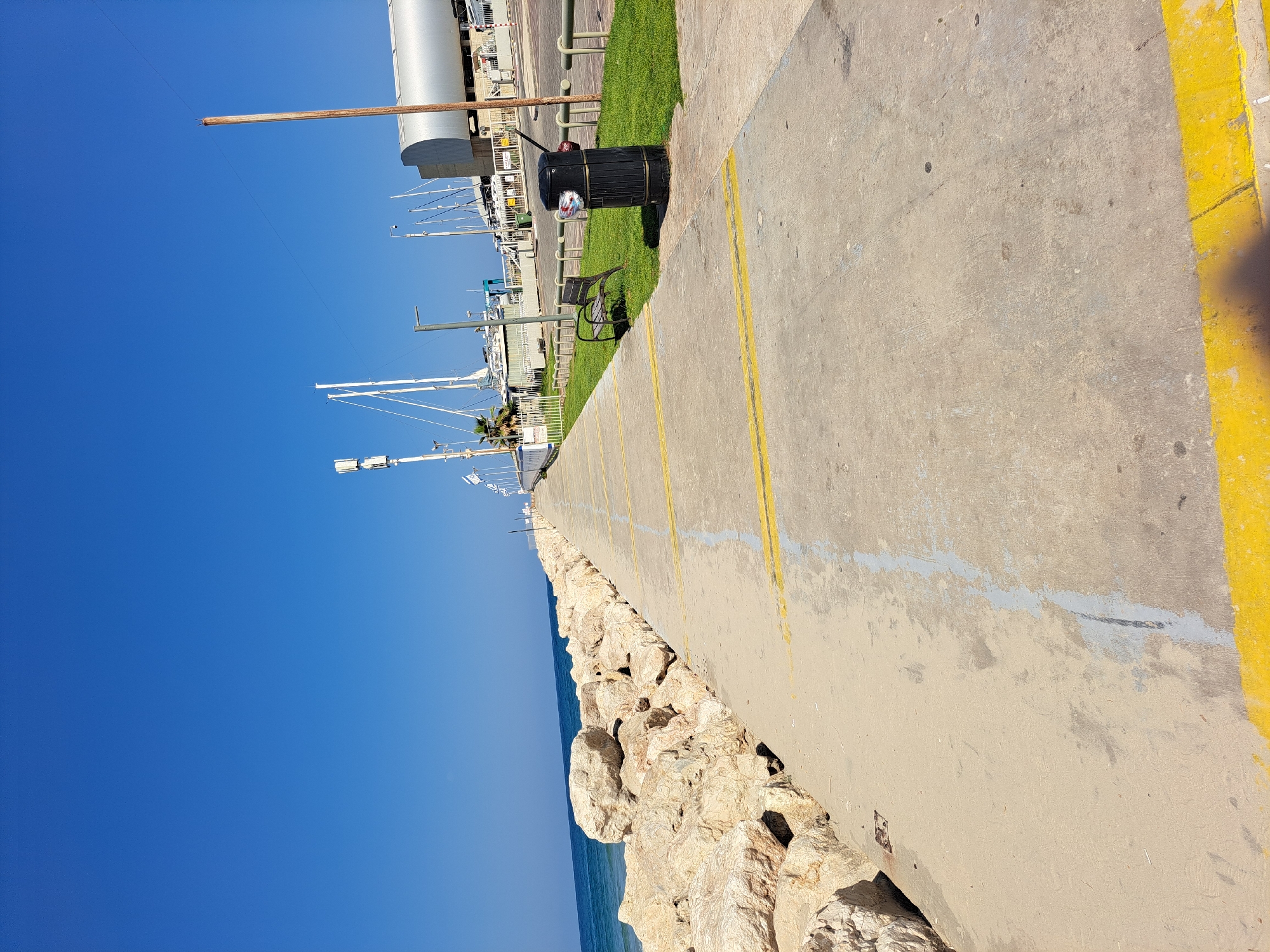 The image depicts a concrete walkway alongside a rocky breakwater on the coast of Herzliya, Israel. The walkway is marked with yellow lines and has a few faded grey lines. To the left of the walkway, large rocks form a barrier between the path and the sea, which appears calm under a clear blue sky. On the right side of the walkway, there is a strip of green grass, a black trash can, a metal bench, and a series of metal railings. In the background, several sailboats are docked at a marina, with buildings and what appear to be communication towers visible. A wooden pole stands vertically on the right side of the frame, near a building. The scene appears to be a public promenade, likely part of the Herzliya marina or its adjacent coastline.
