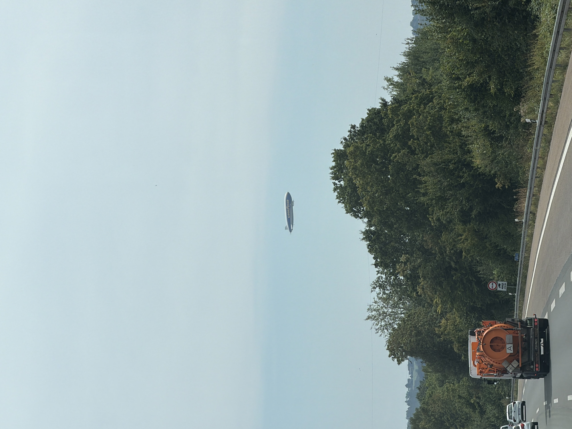 The image depicts a Goodyear blimp flying over a highway in a rural area, with dense green trees lining the side of the road. The blimp is white with blue lettering that reads "GOODYEAR". A large orange truck with a cylindrical tank is visible on the highway in the foreground. Several other vehicles are also present on the highway. The sky is a pale blue, with some hazy atmospheric conditions. The scene appears to be captured during daylight hours. The image does not provide any specific clues to pinpoint the exact location within Dortmund, Germany.