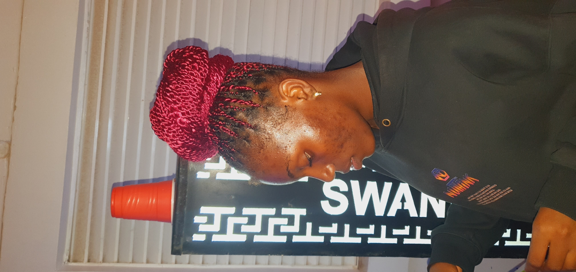 A young Black woman with dark skin and braided hair styled in a large bun on top of her head is shown from the chest up, in profile, facing left. She is wearing a black hooded sweatshirt with a colorful logo on the left chest that appears to read "NUHINK" in iridescent lettering. She has a small gold hoop earring in her left ear. Her skin is shiny, possibly from sweat or lotion, and she has some acne scarring on her cheeks. Her eyes are closed and her lips are slightly parted.
Behind her, to her left, is a tall, dark, rectangular object with a white illuminated geometric pattern on its front. The letters "SWAN" are visible in large white text vertically on this object, and above that, a smaller vertical line of text is partially visible. On top of this object, to the woman's left, is a bright orange plastic cup with horizontal ridges. The background behind the woman consists of horizontal blinds. The scene appears to be indoors, possibly in a dimly lit room. The provided metadata indicates the location is Gura Top, Nigeria.