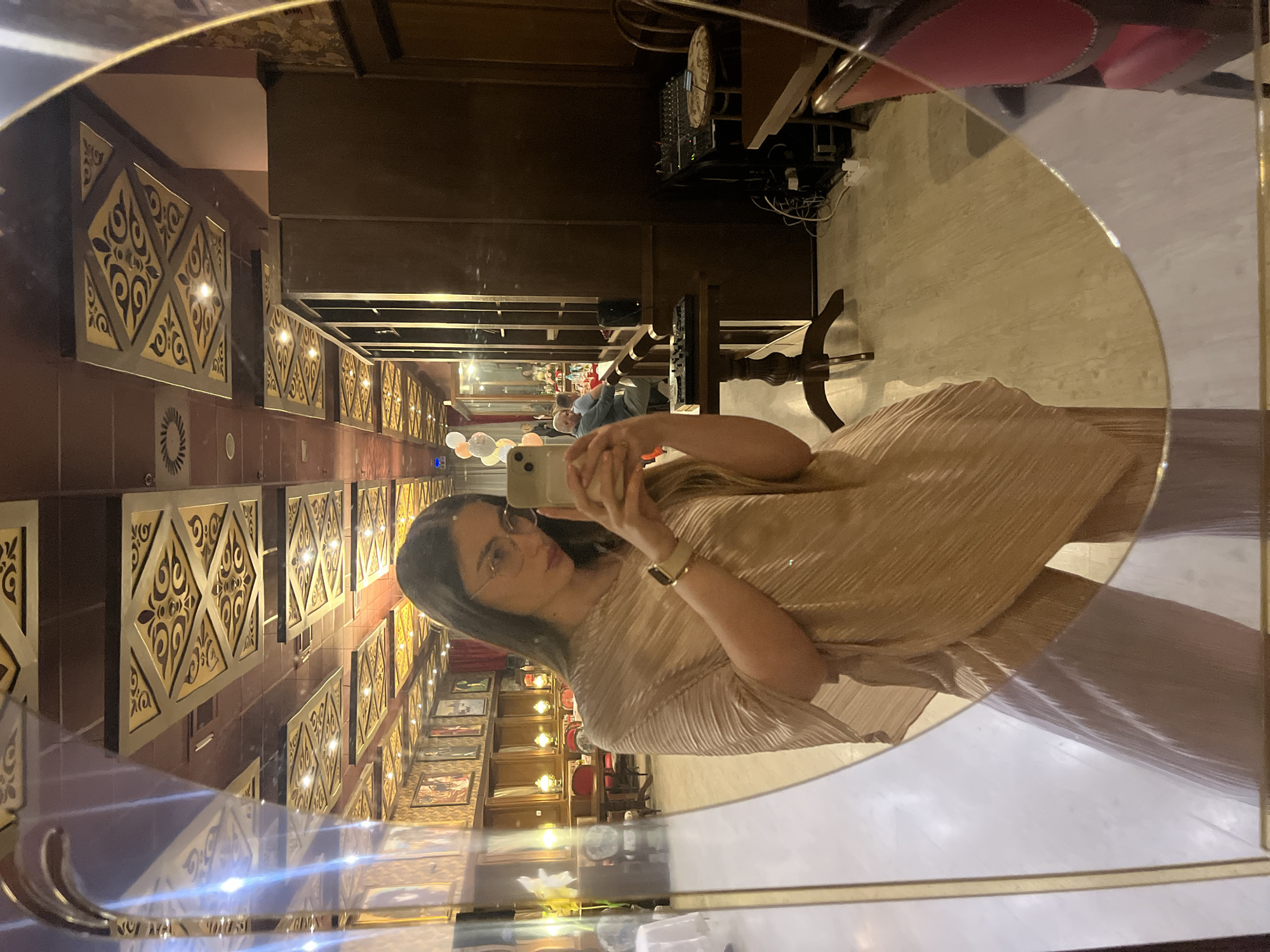A woman is taking a selfie in a round mirror, reflecting the interior of a building. The mirror's frame is gold. The woman has dark hair, wears glasses, and a gold dress or top. She holds a cell phone in front of her face to take the picture, and a watch is visible on her wrist.
The interior features a long room with a decorative wall lined with panels containing ornate designs in gold and dark colors. Further down the hall are pictures and other decor. The room appears to be a restaurant or bar, with tables and chairs visible, along with other patrons at the end of the room. The ceiling has dark wooden beams. There is equipment (perhaps for sound) located on a platform in the background. The lighting is warm and ambient.
The location appears to be a dining establishment. The city and specific location are Unknown, Unknown.