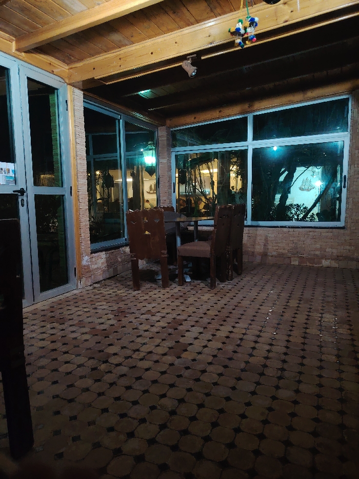 The scene depicts an unoccupied, semi-enclosed patio or veranda in Tanger, Morocco, observed at night. The area features a ceiling constructed from wooden planks and beams. Large, light-framed glass windows and a glass door form the perimeter, with vertical and lower wall sections clad in light brown brick-like tiles. The floor is tiled with a pattern of terracotta-colored octagonal tiles interspersed with smaller dark square tiles.
In the center, a dark-topped round table is surrounded by three dark-stained wooden chairs with carved backs and upholstered seats. A multicolored pompom decoration hangs from the ceiling, near a track light fixture. Through the windows, a dark exterior environment is visible, including the silhouette of a large tree and a faint, illuminated outline resembling a sailing ship on the far right. An interior glass partition reveals an adjacent, internally lit space featuring a traditional green lantern. The overall lighting is dim, with reflections from internal and external light sources visible on the glass surfaces. No individuals are present in the image.