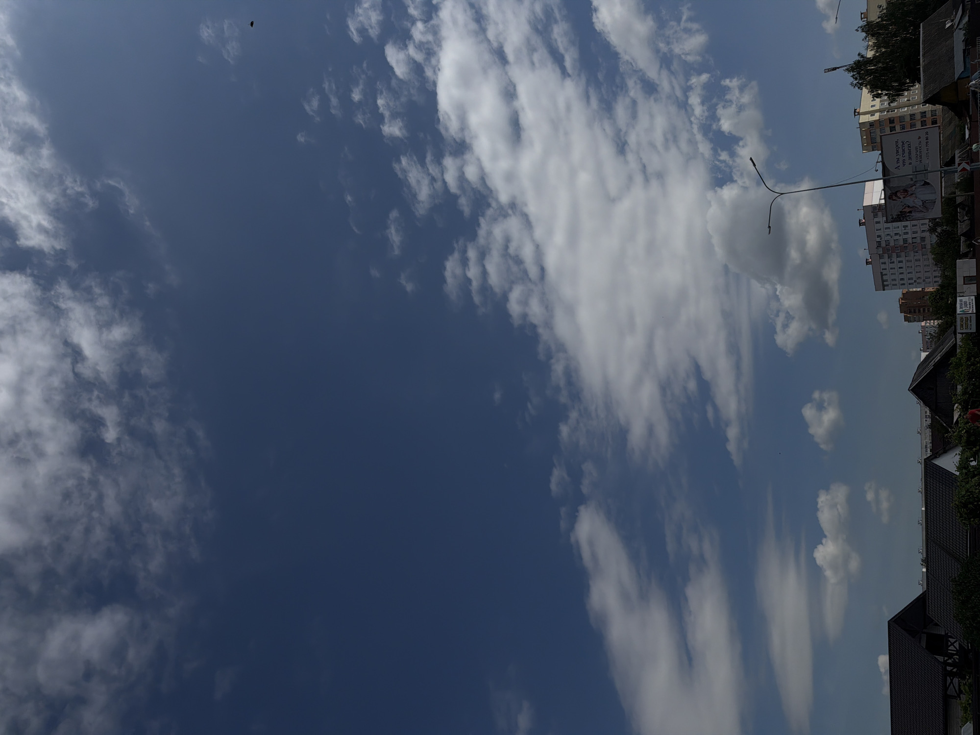 The image shows a wide, upward-facing view of a bright blue sky filled with fluffy white clouds. In the lower portion of the image, a cityscape is visible, featuring the rooftops of several buildings, including some houses with dark, sloped roofs and several multi-story apartment buildings. A streetlamp is prominent in the mid-ground, and a billboard with text and an image is attached to one of the buildings. Some trees and other vegetation are visible between the buildings. A small, dark object, possibly a bird or a drone, is visible against the sky in the upper right quadrant. The overall scene suggests an outdoor environment during daytime, likely in a developed urban or suburban area. The presence of the specific apartment building styles and the Cyrillic text on the billboard point to a location in Belarus. Based on the context of similar images and architectural styles, this scene is likely from Mahilyow, Belarus.