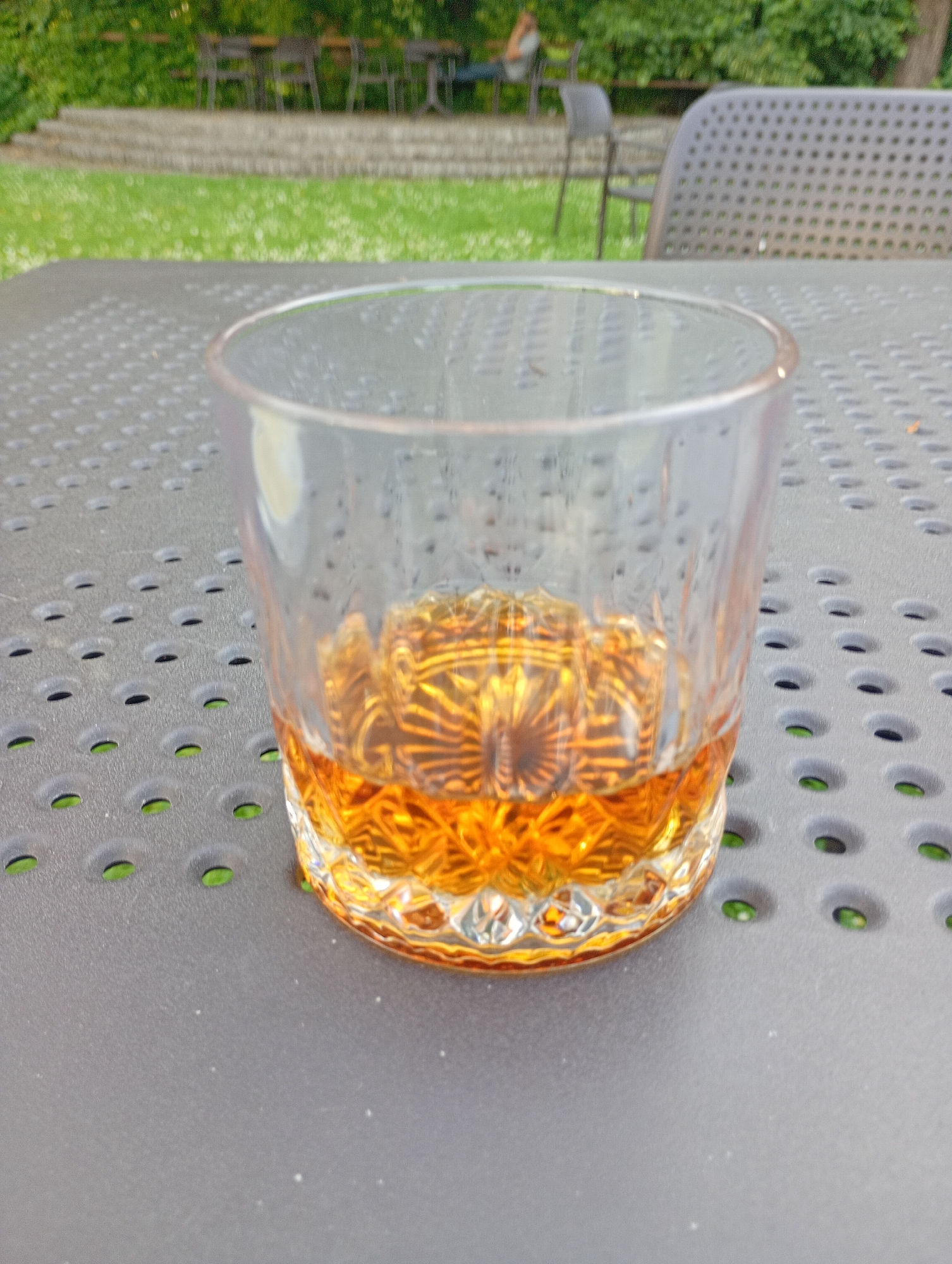 A close-up, eye-level shot shows a clear glass filled with amber liquid, likely whiskey or brandy, resting on a gray, perforated outdoor table. The glass has a textured, faceted design at the bottom, and condensation is visible on its exterior. The liquid reflects the patterned glass and light.
In the background, out of focus, are elements of an outdoor seating area and garden. A green lawn with scattered white flowers is visible behind the table. Further back, there are stone steps leading up to an area with several dark chairs and tables, suggesting a patio or garden seating arrangement. Beyond that, lush green foliage forms a backdrop. To the right, a portion of a dark, perforated outdoor chair or lounge is visible.
The scene appears to be set in a garden or patio area, possibly a restaurant or beer garden. The style of the furniture and the presence of a well-maintained lawn are consistent with an outdoor seating area in a public space or a private residence. The lighting suggests it is daytime, possibly late afternoon or early evening, given the warm tones of the liquid and the soft background lighting. The overall impression is one of a relaxed outdoor setting. The location is in Międzyrzecz, Poland.