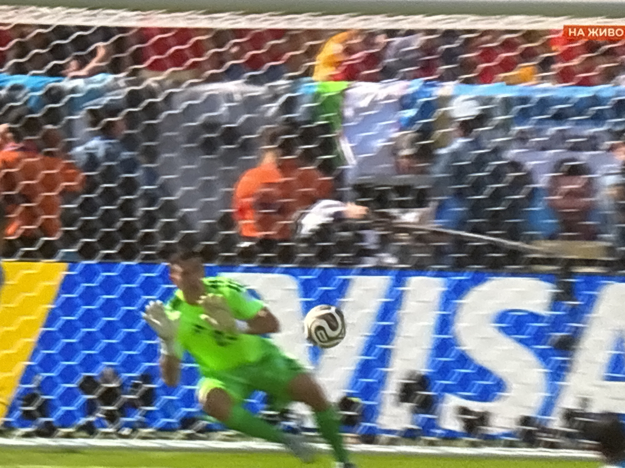 The image shows a blurry action shot from a soccer match. A goalkeeper, dressed in a bright green uniform, is in the process of diving to save a soccer ball, which is captured mid-air. The background reveals a crowd of spectators behind a net, with some individuals wearing orange and red. The scene appears to be outdoors, with a large blue and white banner featuring the word "VISA" visible behind the goal. A small red rectangle with white text "НА ЖИВО" (meaning "Live" in Bulgarian) is superimposed on the upper right corner of the image. The image's overall quality is degraded by motion blur and pixelation, making it difficult to discern precise details. The mention of Burgas, Bulgaria, is not supported by any visual evidence within the image itself.