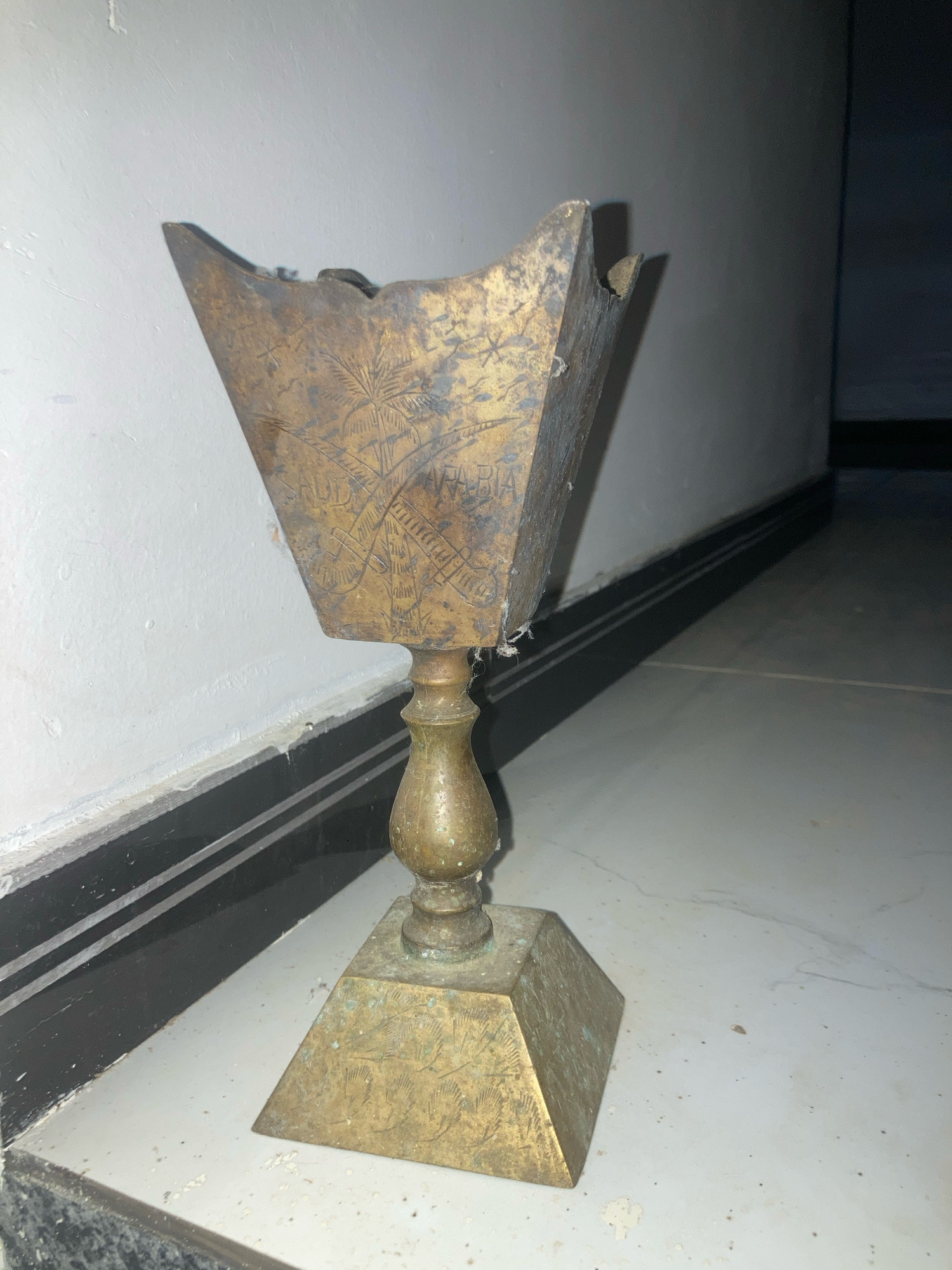 An antique brass incense burner, likely used for burning bakhoor or similar aromatic substances, stands on a tiled floor. The burner features intricate etchings of palm trees and other decorative motifs on its conical top and square base. The metal exhibits signs of age and wear, with patina and some damage visible on the upper rim. The burner is positioned in a dimly lit interior, with a white wall and a black and white striped skirting board forming the backdrop. The floor tiles are light-colored and show some dust and debris, suggesting a storage or less-frequented area. The lighting casts shadows, highlighting the textures and details of the object. The location is identified as Daladagama, Sri Lanka.
