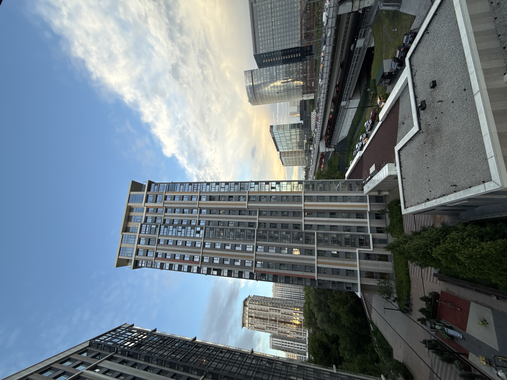 The image depicts a cityscape in Khimki, Russia. It shows several modern high-rise buildings with glass and concrete facades under a sky with blue patches and scattered white clouds. An elevated highway with vehicular traffic runs through the scene. Lower in the frame, trees and walkways are visible, suggesting a park or residential area. The overall lighting indicates either dawn or dusk.