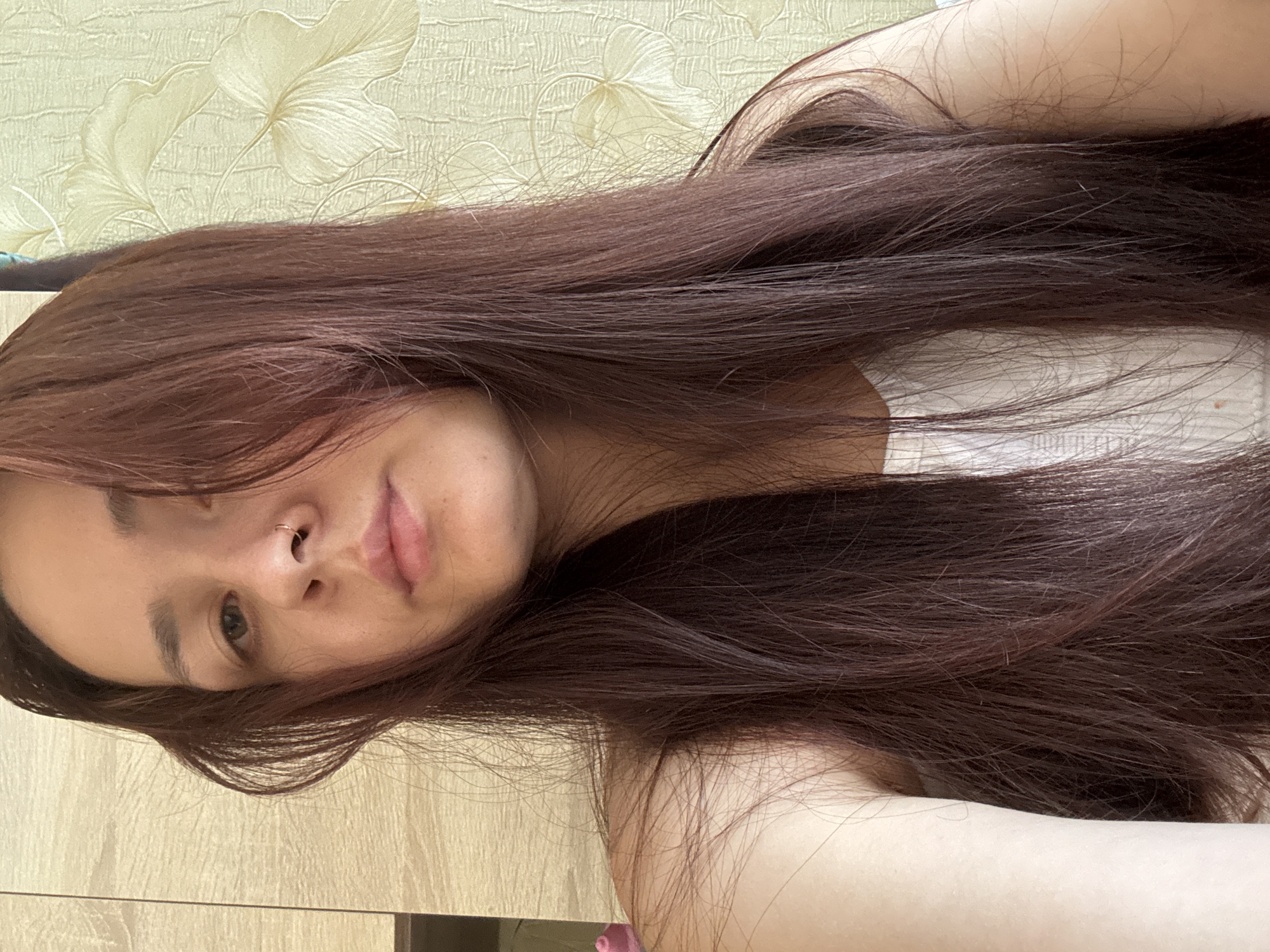 The image shows a young woman with long, dark brown hair, wearing a white top. She is looking towards the camera, with her hair falling over her shoulders. Her face is partially obscured by her hair. In the background, there is a wall with a light-colored floral pattern, and to the left, a light-colored wooden cabinet or wall. The lighting suggests an indoor environment. The image was taken in Naberezhnye Chelny, Russia.