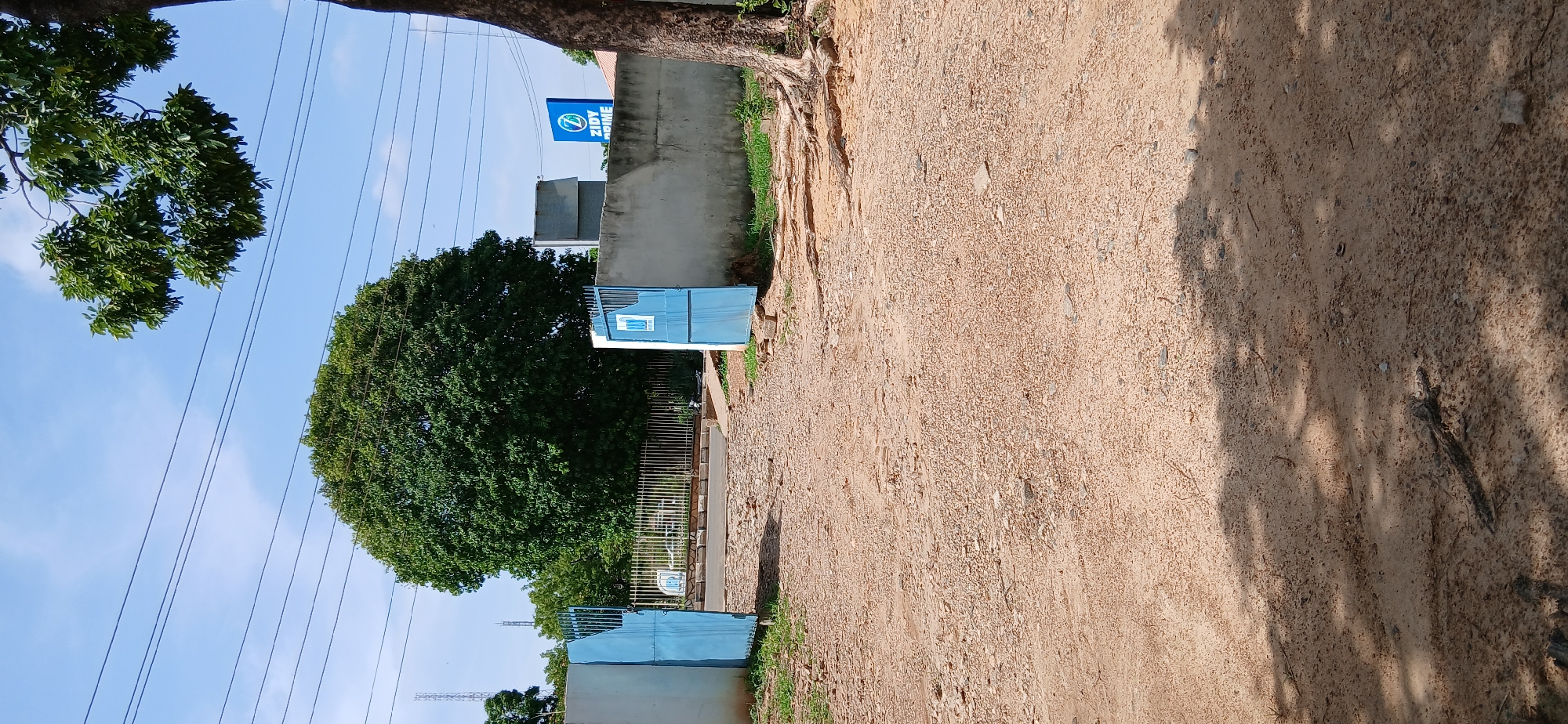 The image captures a bright, sunny outdoor scene, likely a roadside or path in Shaba, Nigeria. The dominant feature on the right side is a wide, light brown dirt path, heavily textured with sand and small stones. Shadows from unseen foliage above are cast across this path, indicating strong sunlight. Along the immediate edge of the path on the left, exposed tree roots are visible, transitioning into a grassy area.
On the left side of the frame, several structures and natural elements are present. A large, verdant tree with dense green foliage stands prominently. Further along, a concrete wall or fence line runs parallel to the path. Above this wall, a rectangular blue sign displays a white circular logo, possibly a stylized "Z" or "S," next to the legible white text "ZIDY HOME."
Below the sign, two light blue (or teal) metal structures, resembling gates or small enclosures, are visible. The closer one features a small white rectangular detail. Between these blue structures, a section of white metal fencing is also present. High above, several thin utility lines or power cables crisscross the clear blue sky, which has a few faint white clouds. The scene is illuminated by bright daylight, suggesting mid-morning to late afternoon. No people or active events are depicted.