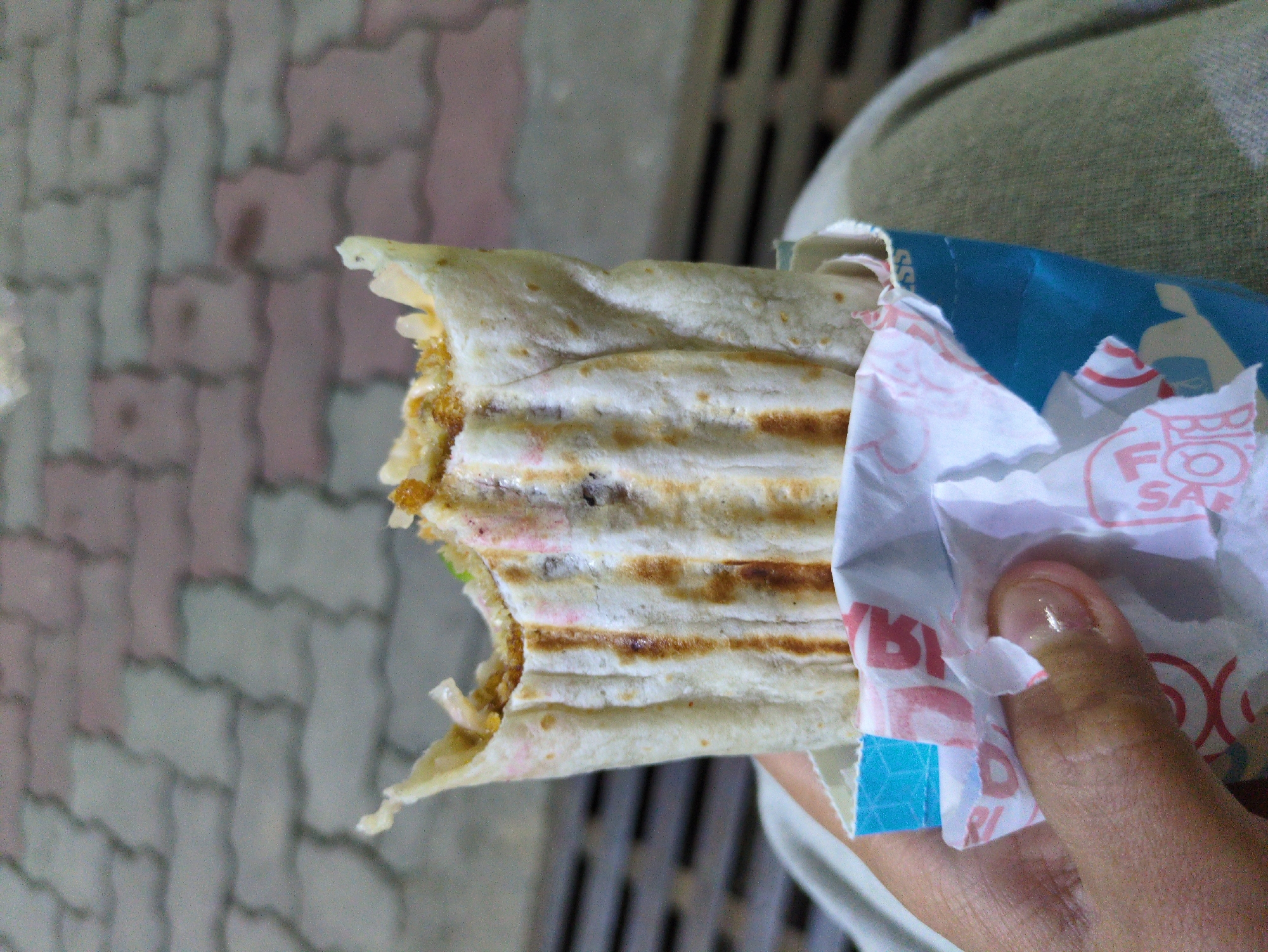A person's hand is holding a partially eaten shawarma, which is wrapped in paper and a blue container. The shawarma has grill marks and appears to contain meat and vegetables. The background shows a brick-like pavement, suggesting an outdoor setting. The person is wearing a light-colored garment, possibly pants or a skirt. The scene is likely from Dharamshala, India, based on the contextual information provided.