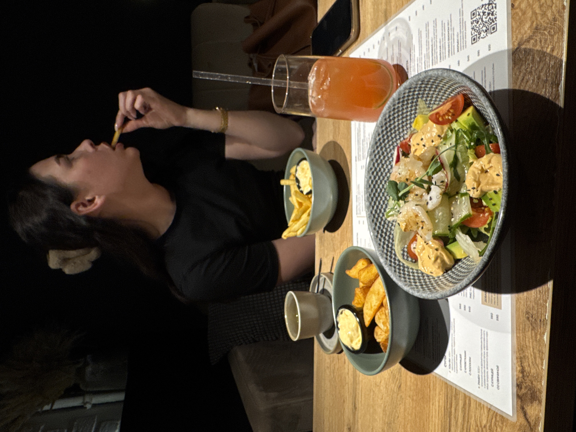 A woman is eating a french fry at a table in a restaurant. The table has a salad, a drink with a straw, two small bowls of french fries and a small container of sauce. A menu is on the table. The location is likely Chaikovsky, Russia, given the menu contains some writing in Russian. The light is low and it is likely evening.