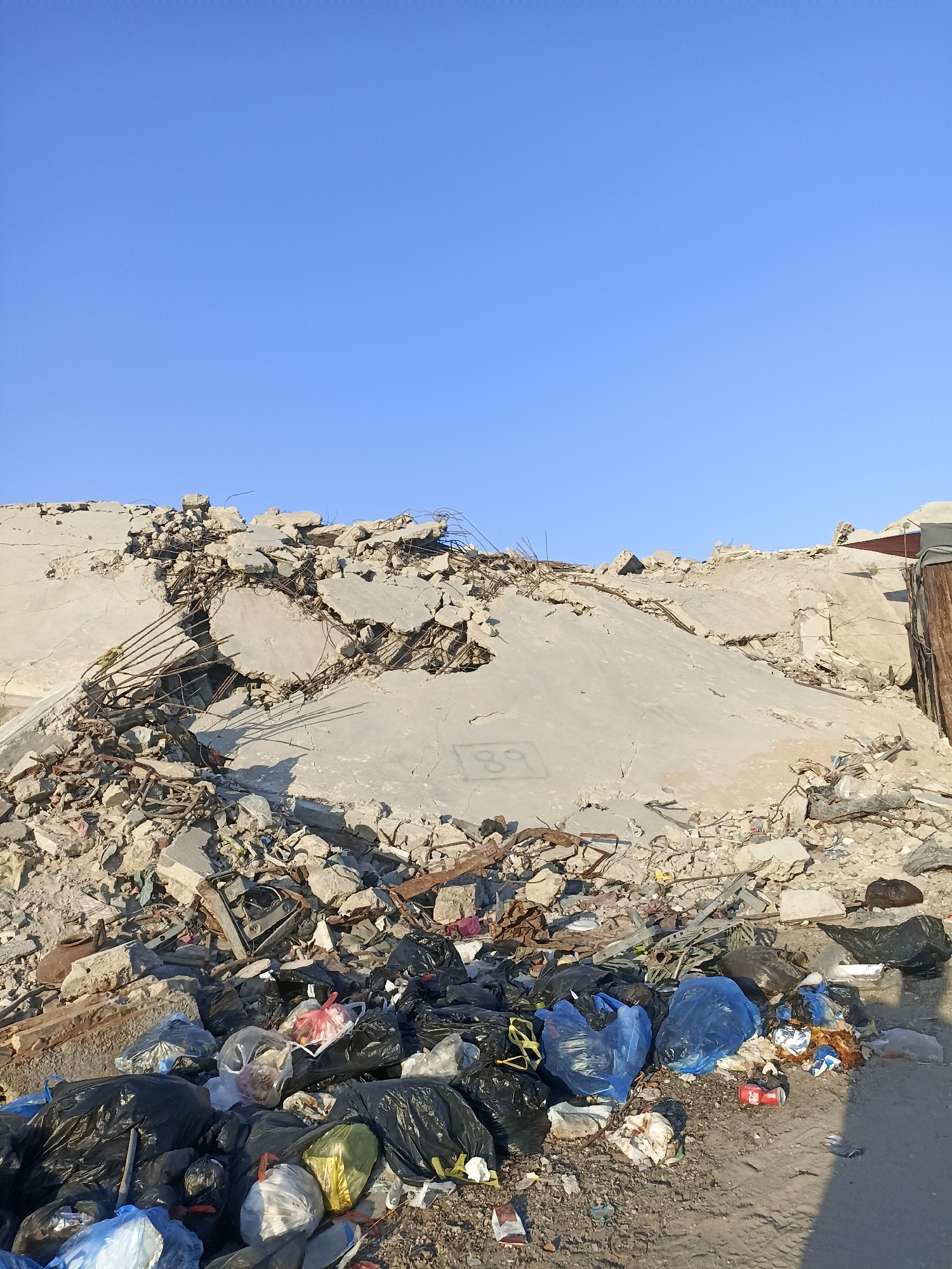 The image shows a pile of rubble from a destroyed building, with scattered debris and trash bags in the foreground. The rubble consists of concrete, rebar, and other building materials. The trash bags are various colors and sizes, some of which are torn open, revealing their contents. The scene appears to be an outdoor location under a clear blue sky. The presence of the rubble suggests recent destruction, possibly due to conflict. There are no people visible in the image. The location is identified as Gaza City, Palestinian Territories, indicating a context of ongoing conflict and humanitarian crisis.