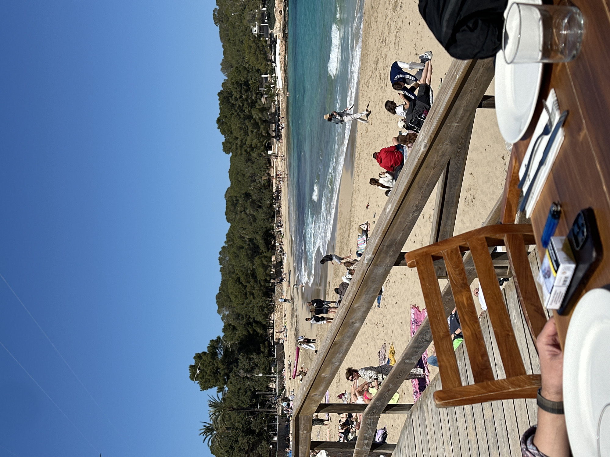 The image shows a beach scene in Sant Josep de sa Talaia, Spain, viewed from a raised wooden deck, possibly a restaurant terrace. The beach is crowded with people sunbathing, swimming, and walking along the shoreline. Turquoise water meets the sandy beach, with a line of trees along the coast dividing the beach from the clear blue sky. In the foreground, a wooden table is set with plates, cutlery, and a glass. A wooden chair is positioned on the deck. A pack of cigarettes and a lighter are on the table near a person's hand with a watch. The scene suggests a relaxed, sunny day at the beach.