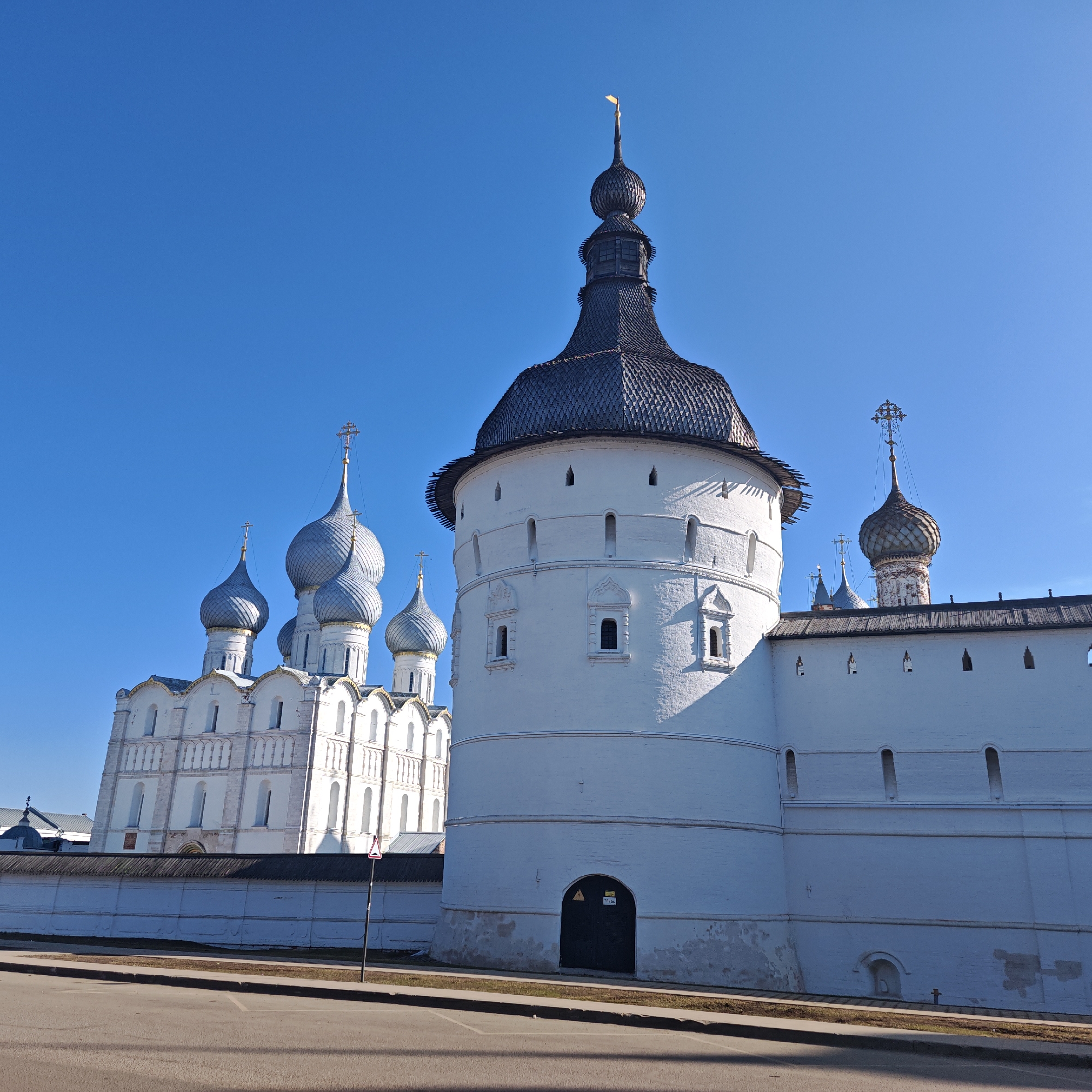 The image shows a bright, sunny day in what is likely Rostov, Russia. A white brick wall and tower of a monastery stand in the foreground. The tower is cylindrical with a dark, shingled roof and a finial at its peak. A dark arched doorway is visible at the base of the tower. Behind the wall and tower is a white building with multiple silver onion domes topped with crosses. A road and sidewalk are in the immediate foreground. A small traffic sign is beside the road. The sky is clear and blue. There are no people visible.