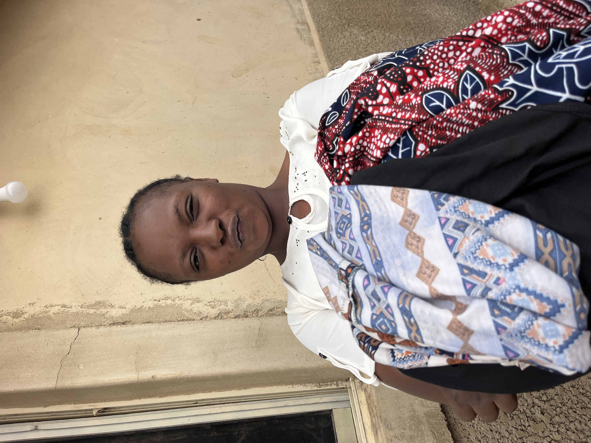 The media file shows a woman from the chest up, holding fabric. The woman is facing the camera and has dark skin and dark hair pulled back from her face. She is wearing a white top with decorative eyelet details around the neckline. She is holding a stack of folded fabric in front of her. The top layer of fabric has a bold pattern in red, white, and blue, featuring abstract shapes and leaf-like motifs. Beneath that is a black garment, and then another fabric with a geometric pattern in shades of blue, white, and brown. The background appears to be an outdoor setting, with a concrete or stucco wall to the left and a gritty ground surface to the right. The lighting suggests it is daytime. The woman's expression is neutral or slightly questioning. No text is visible in the image.